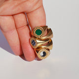 Four signet rings stacked on a finger, all gold toned bronze with different color stones surrounded by a diamond burst pattern