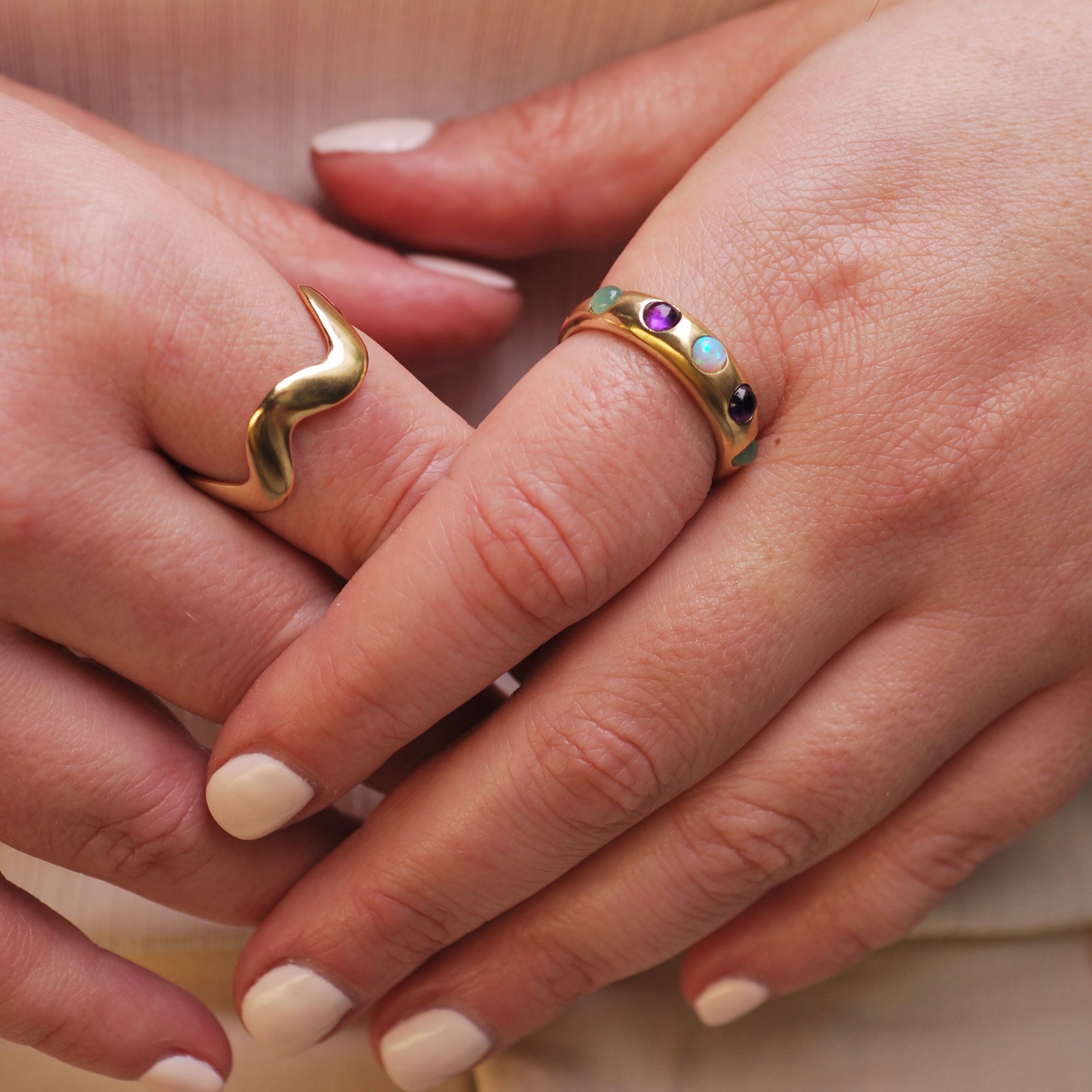 Close-up of two hands wearing gold rings with gemstones on a neutral background