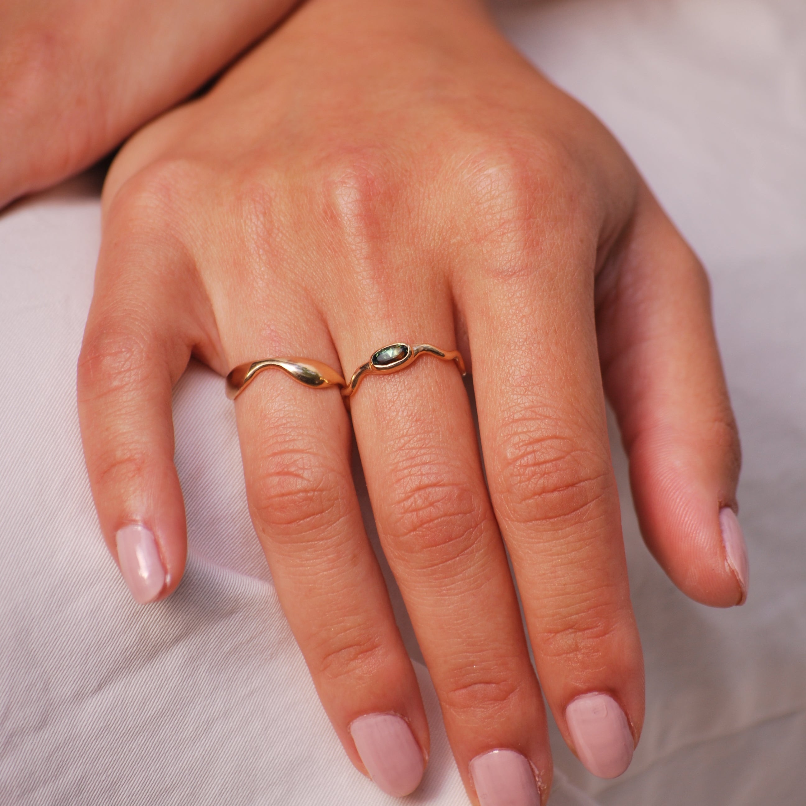 Close-up of a hand wearing two gold wavy organic rings, one with a bi-color sapphire on a light background