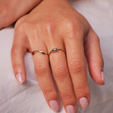 Close-up of a hand wearing two gold wavy organic rings, one with a bi-color sapphire on a light background