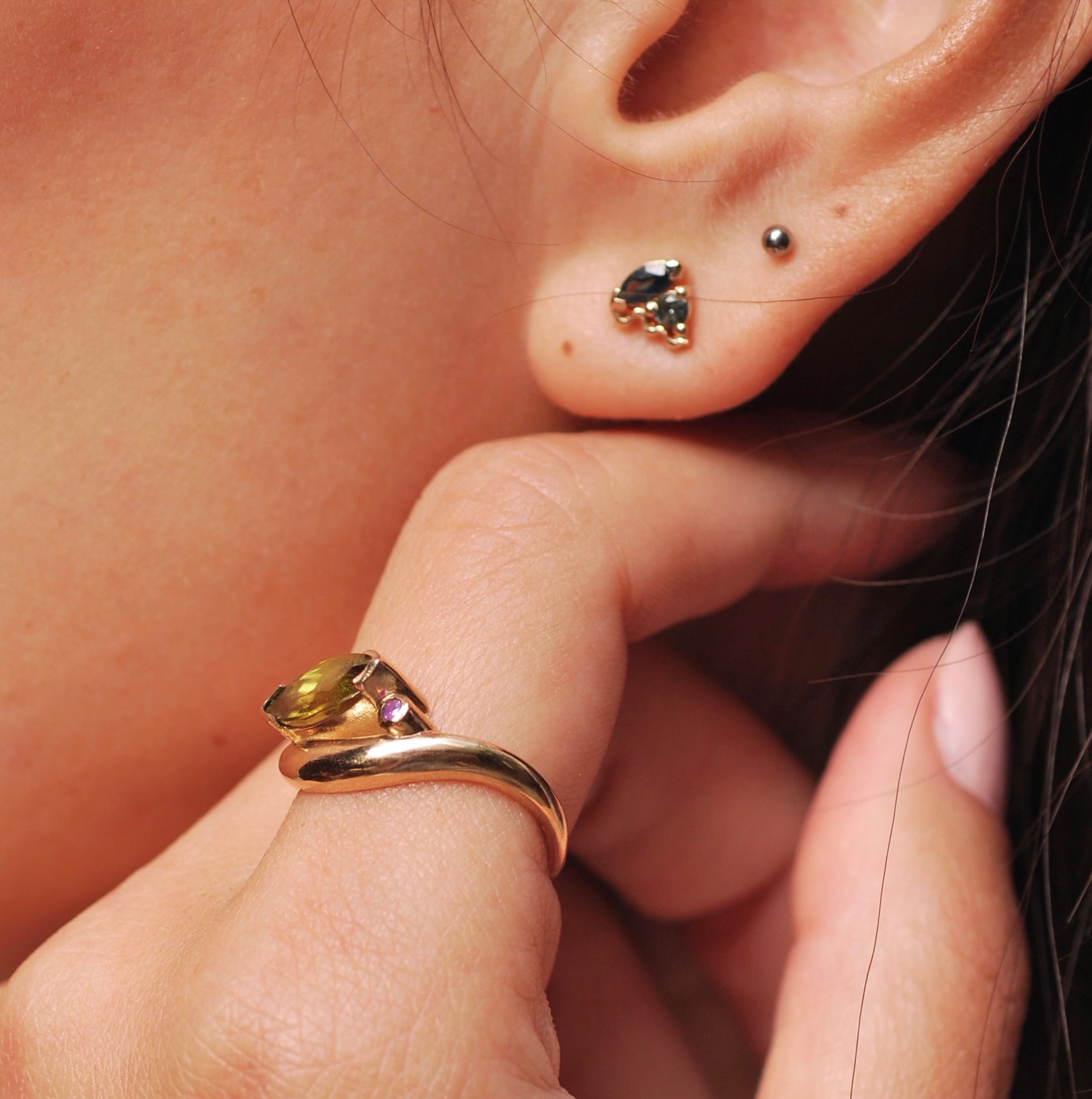 Close-up of an ear with earrings and a hand touching the earlobe. The hand has a side view of a gold ring with green and pink stones.