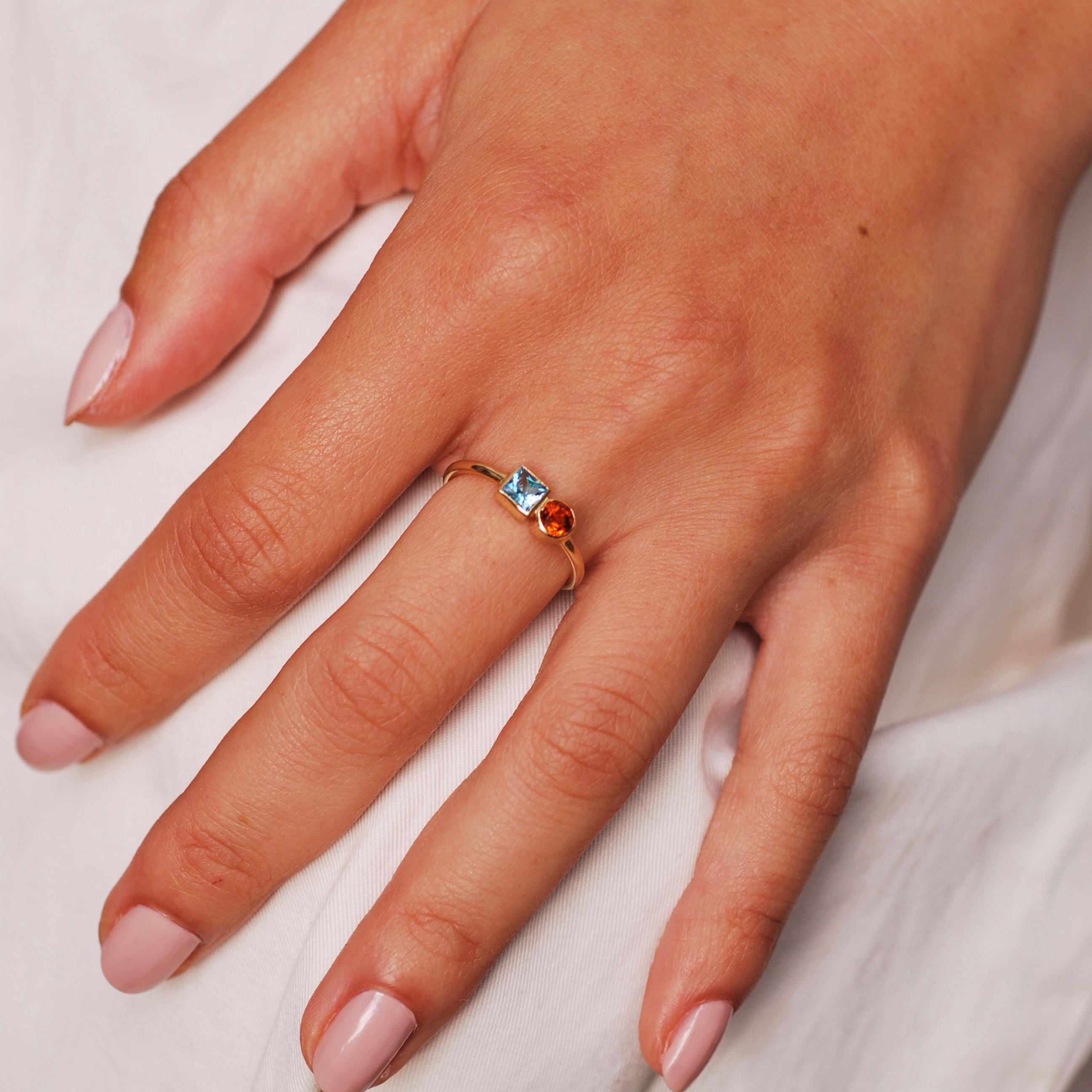 Hand wearing a gold ring with blue and orange gemstones on a light background