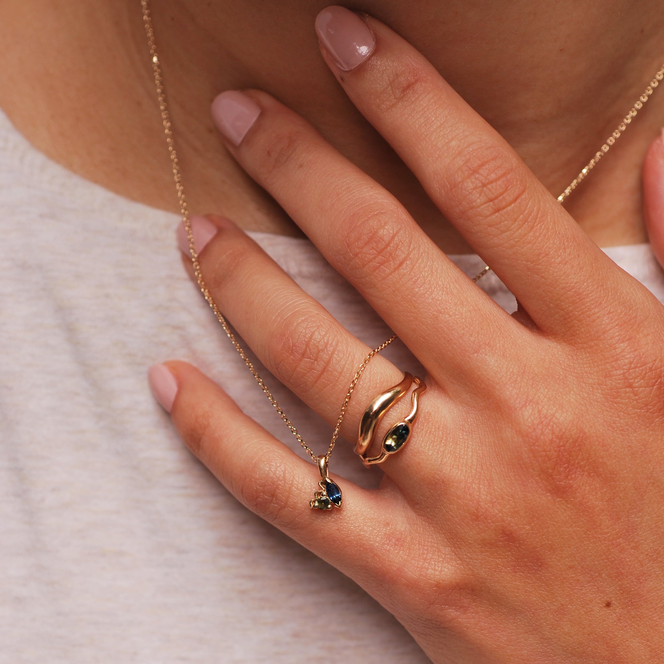 Hand wearing two gold rings, one with an arch and one with a small oval sapphire. Hand is also holding a delicate necklace with a cluster of small blue and green sapphire stones. 