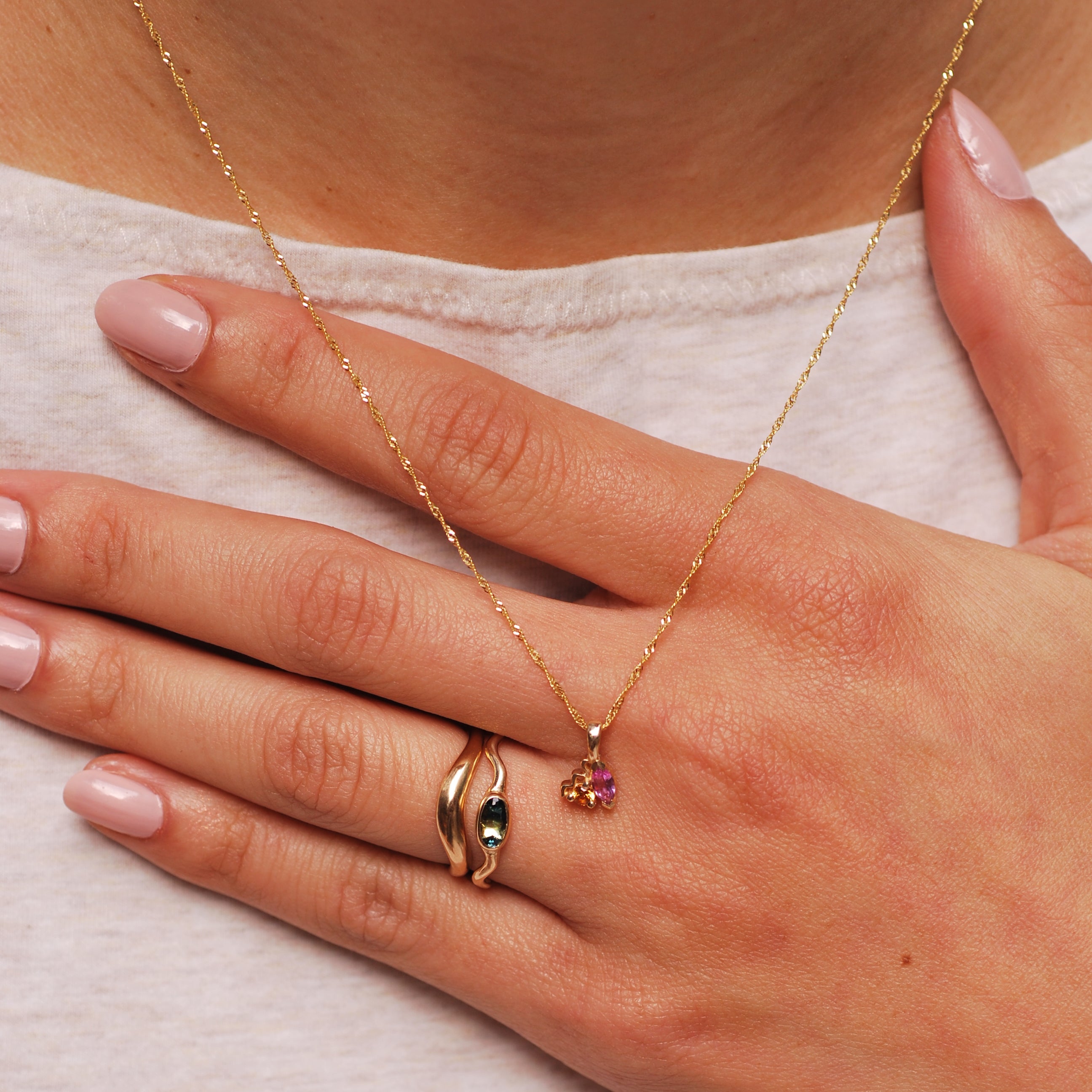 Hand against chest wearing two stacking gold rings, one with an arch and one with a wavy band and an oval bi-color sapphire. A necklace also rests against the hand and is a delicate gold piece with orange and pink stones.