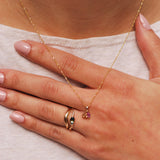 Hand against chest wearing two stacking gold rings, one with an arch and one with a wavy band and an oval bi-color sapphire. A necklace also rests against the hand and is a delicate gold piece with orange and pink stones.