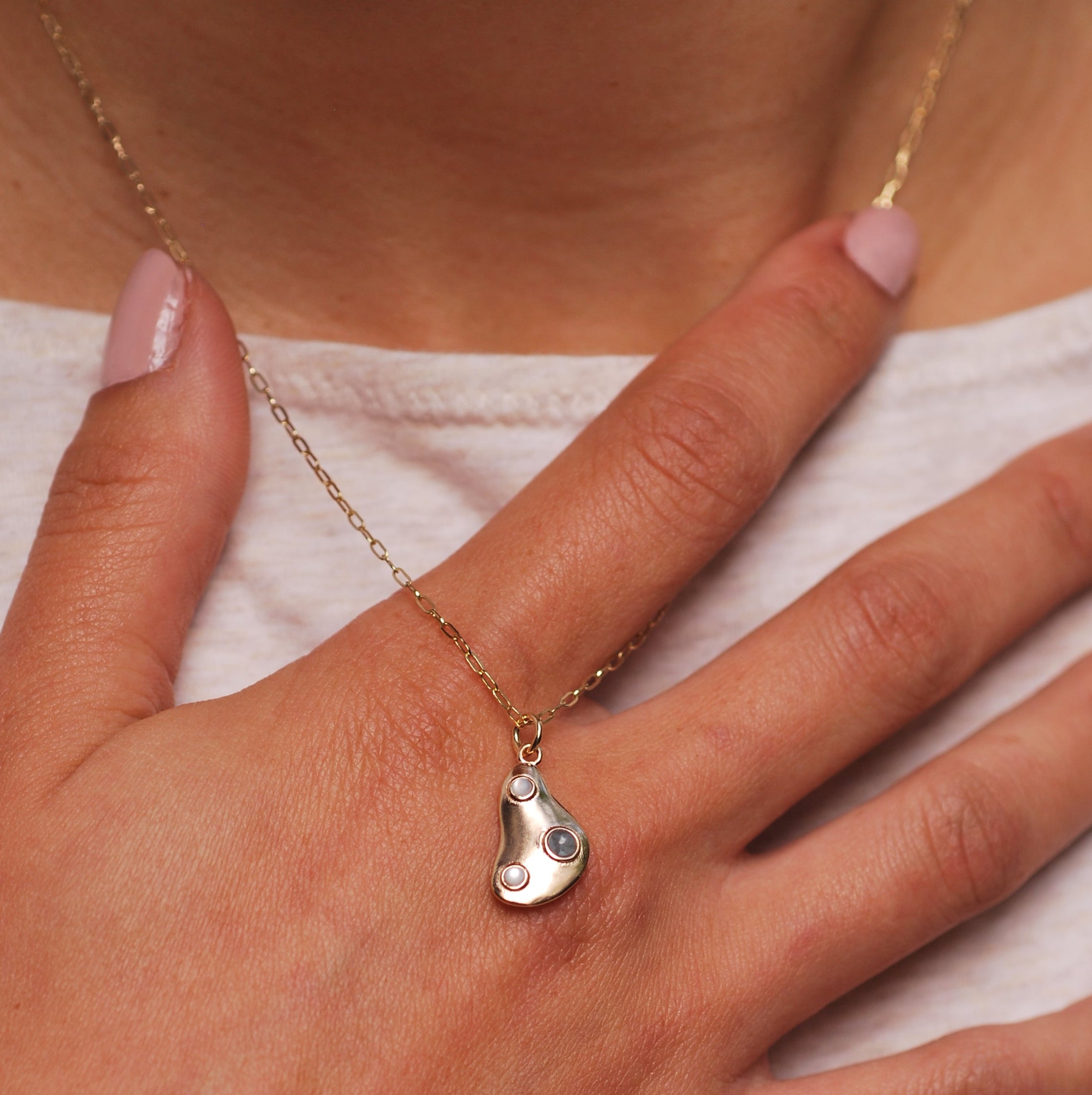 A woman with her hand to her neck with a gold puddle necklace with two mother of pearl and an aquamarine cabochon draping over her fingers. 