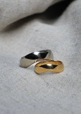 Two hand carved, arched rings, one yellow gold and one white gold, on a textured gray fabric background.