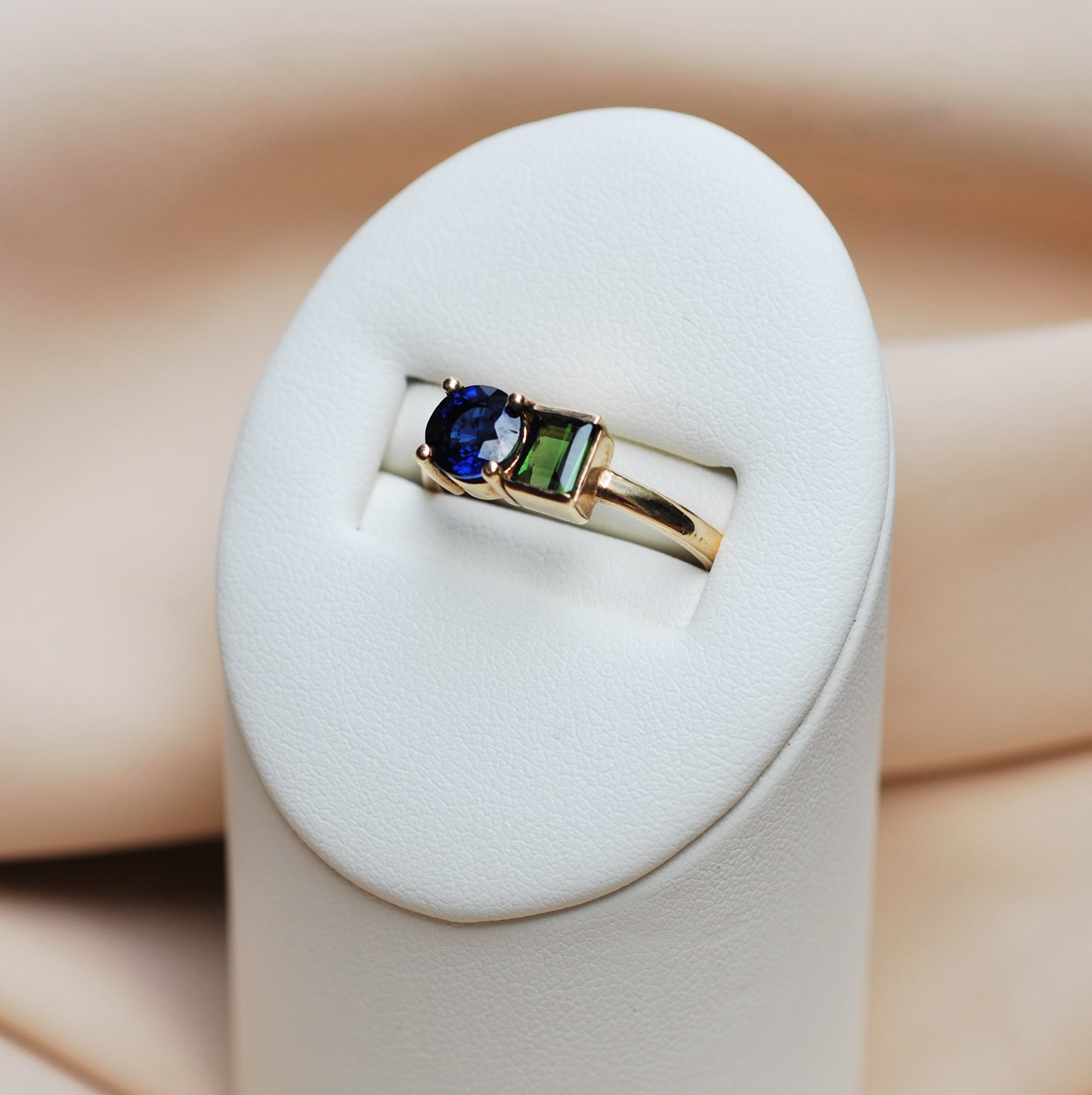 Gold ring with a blue sapphire and green tourmaline on a white stand against a soft pink fabric background