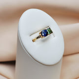Gold ring with a toi et moi setting of a blue sapphire and green tourmaline on a white stand against a beige background