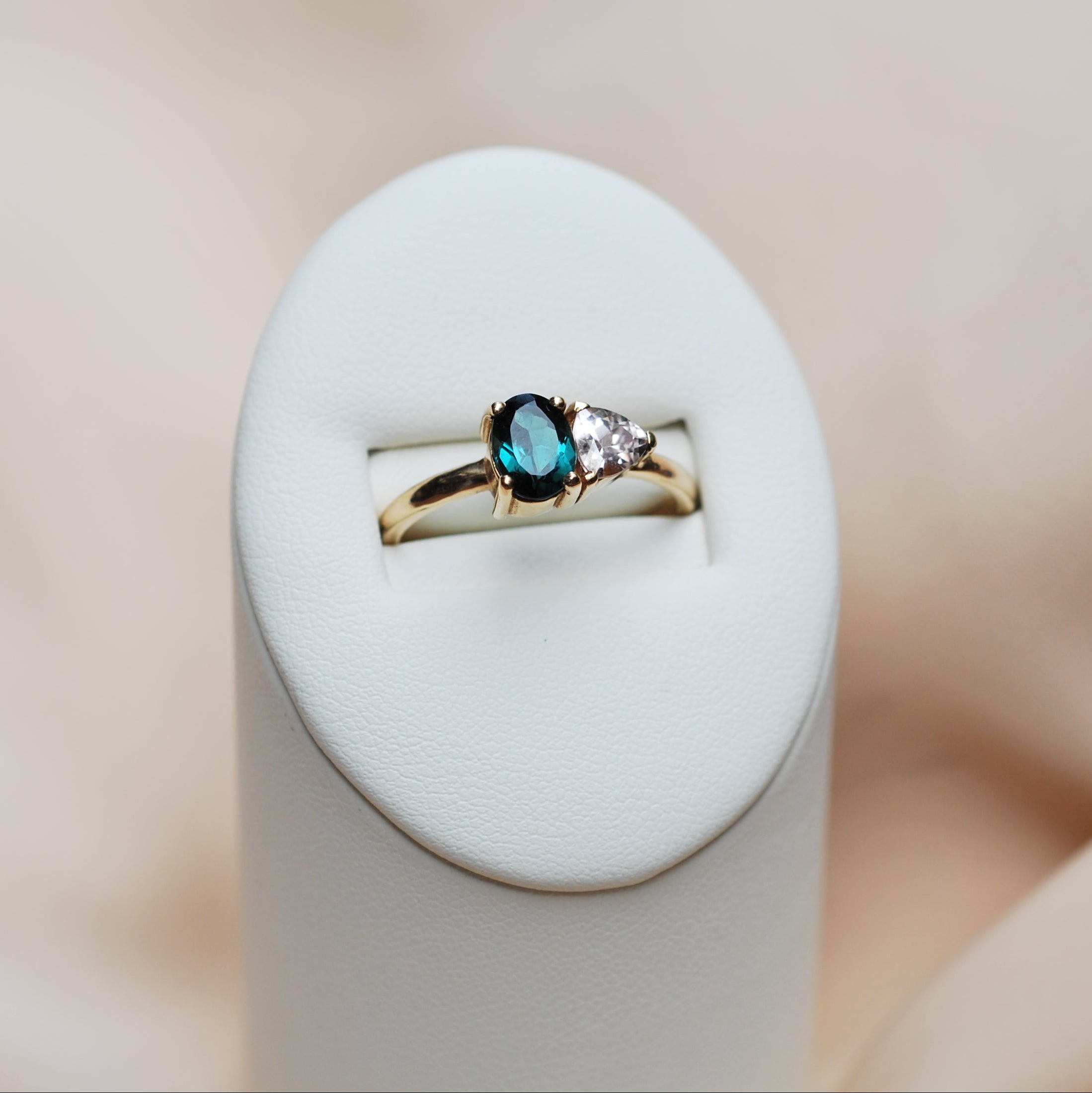 Gold ring with a toi et moi setting of a teal and light pink gemstone on a white stand against a beige background