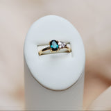 Gold ring with a toi et moi setting of a teal and light pink gemstone on a white stand against a beige background