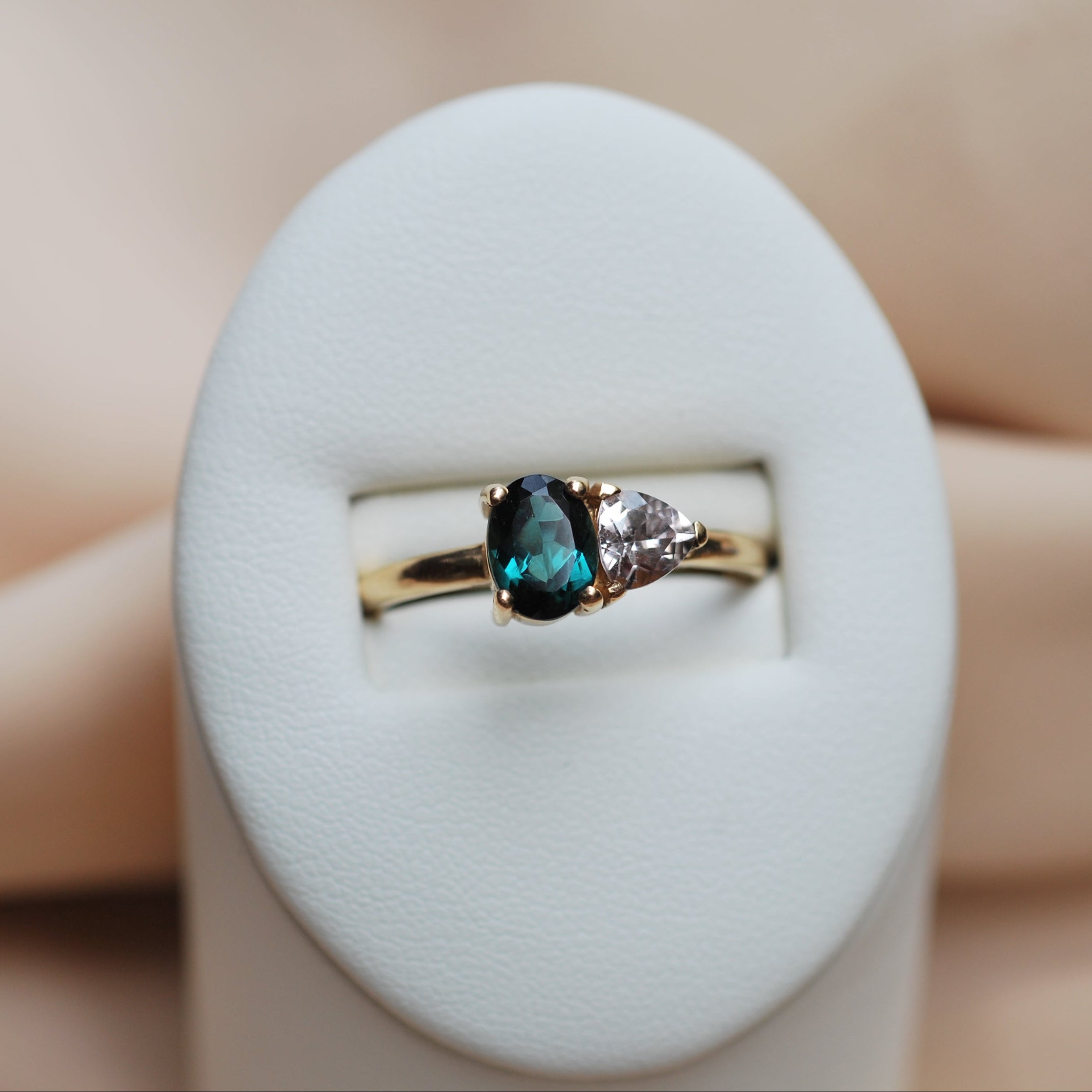 Gold ring with a teal tourmaline and pink sapphire on a white stand against a beige background
