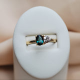 Gold ring with a teal tourmaline and pink sapphire on a white stand against a beige background