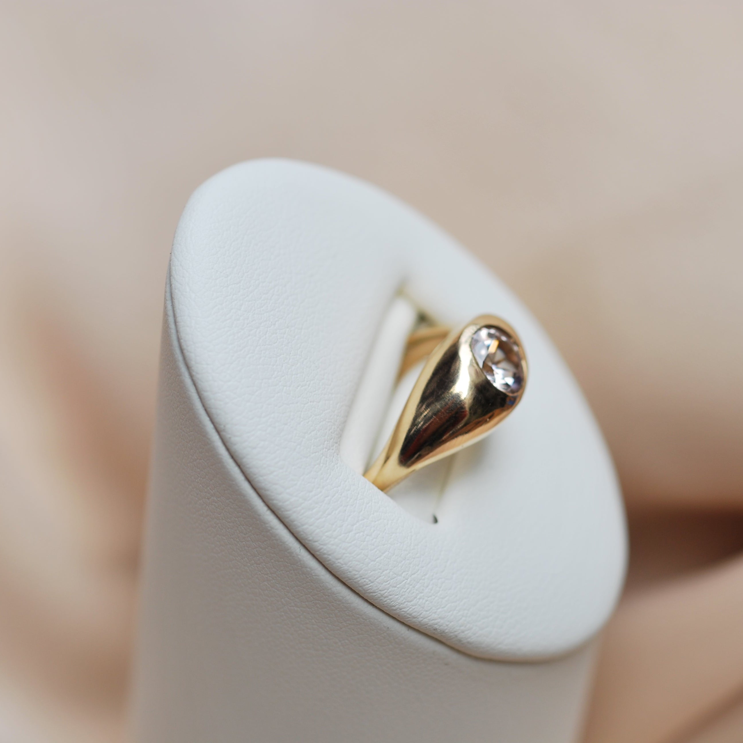 Side view of a gold ring with a flush set zircon on a white stand with a blurred background