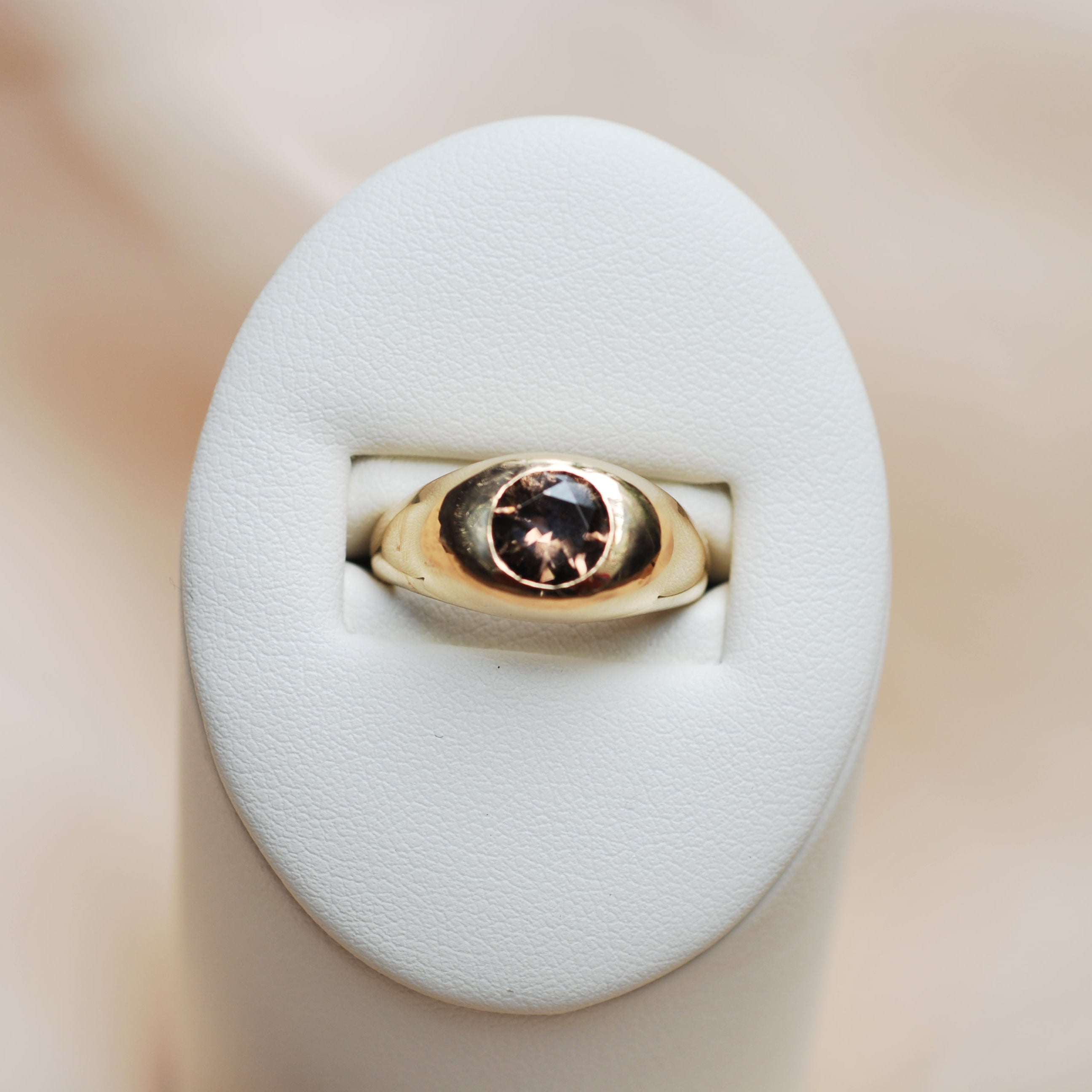 Gold ring with a zircon flush set faceted stone on a white cushion against a beige background