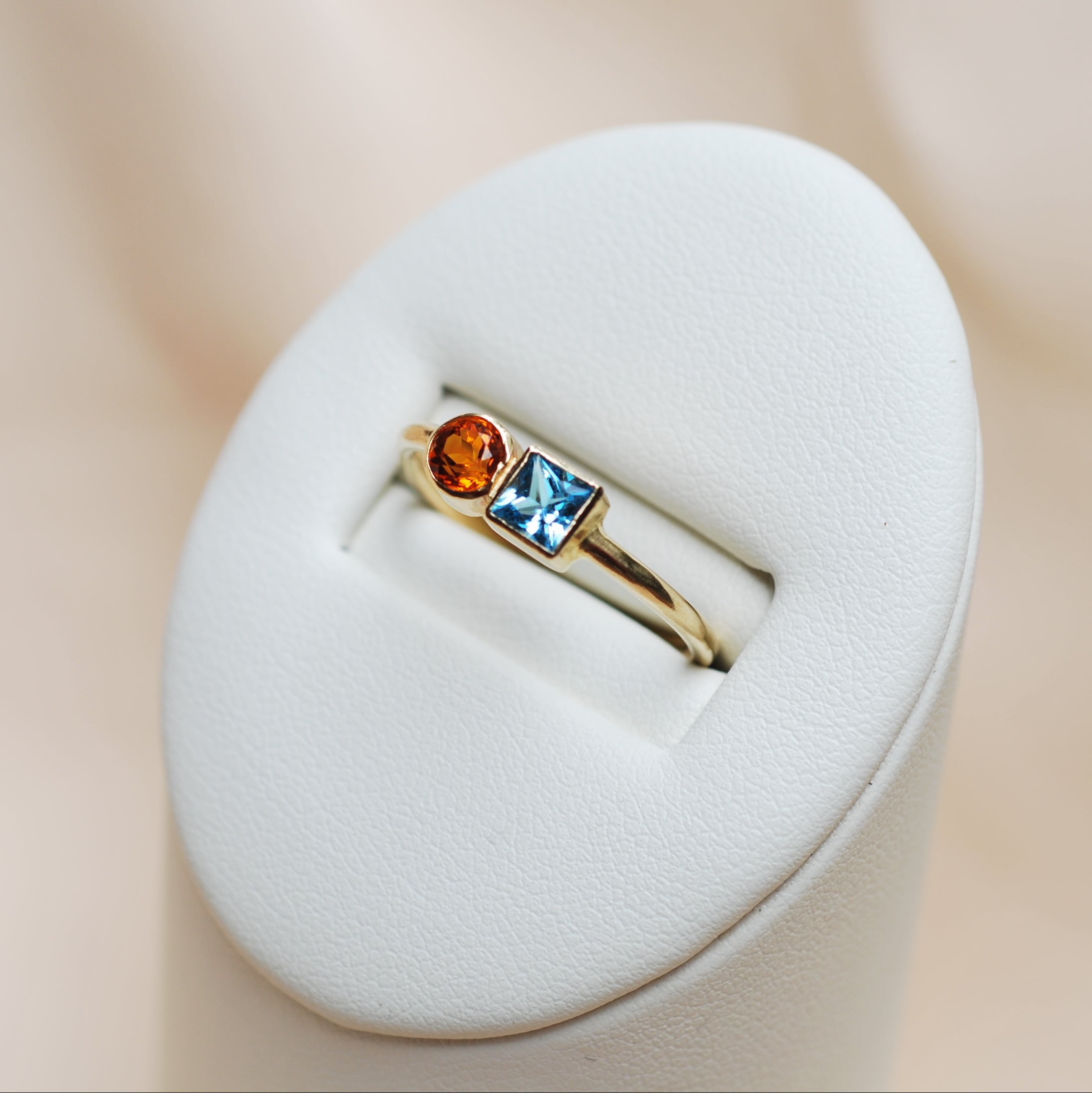 Gold ring with blue and orange gems on a white stand against a beige background