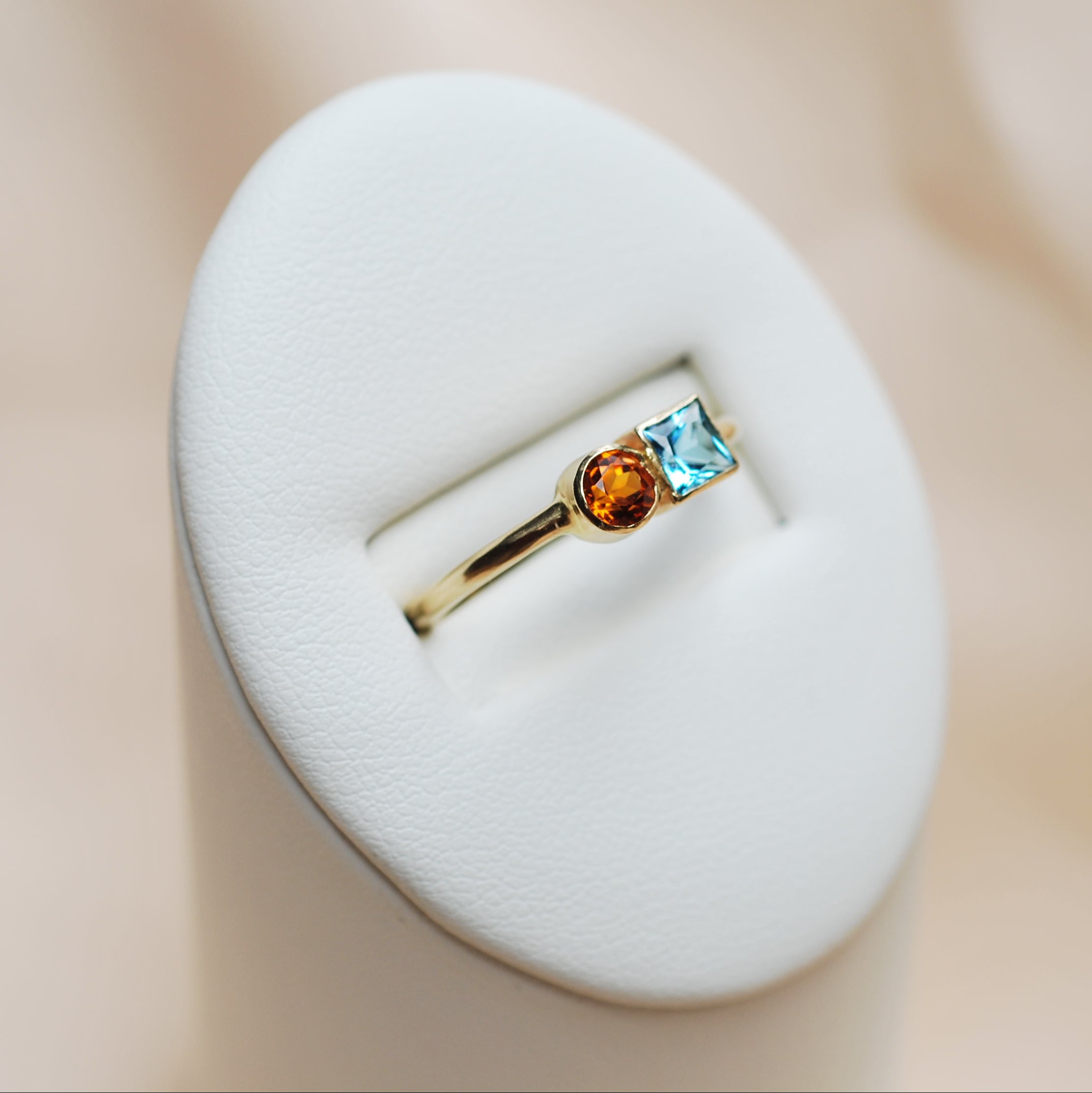 Gold ring with blue and orange gemstones on a white stand against a beige background