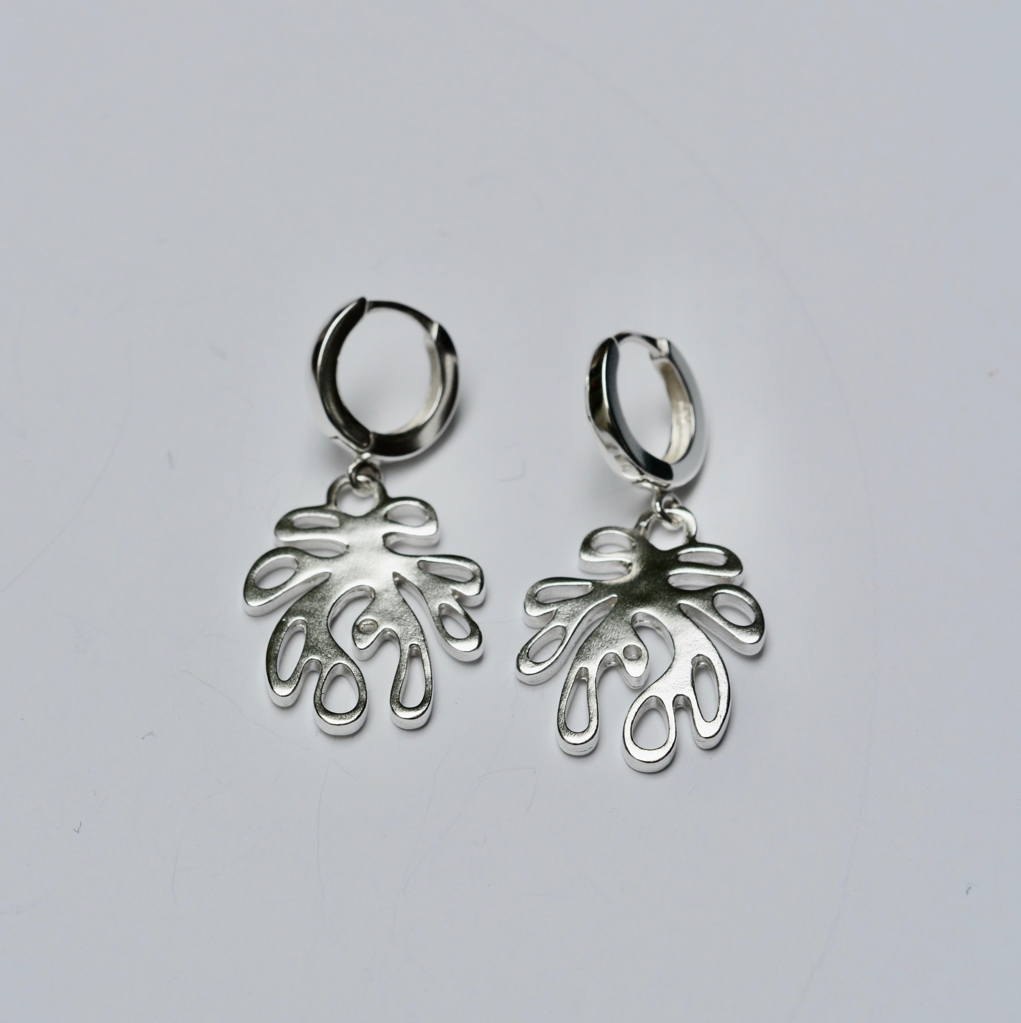 Pair of silver huggie hoop earrings with a coral shaped charm on a light gray background