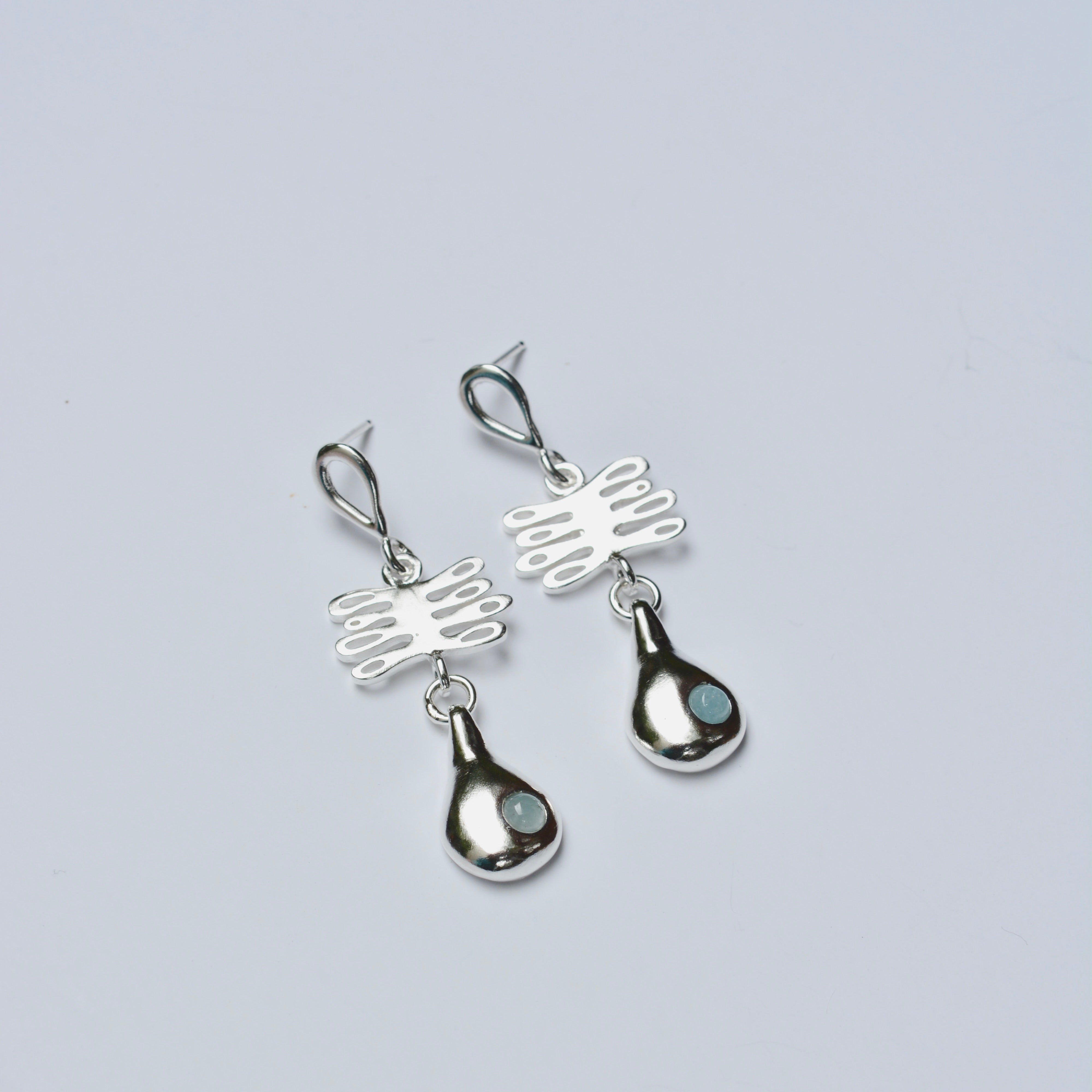 Pair of silver earrings with a plant shaped charm and light blue gemstone on a gray background