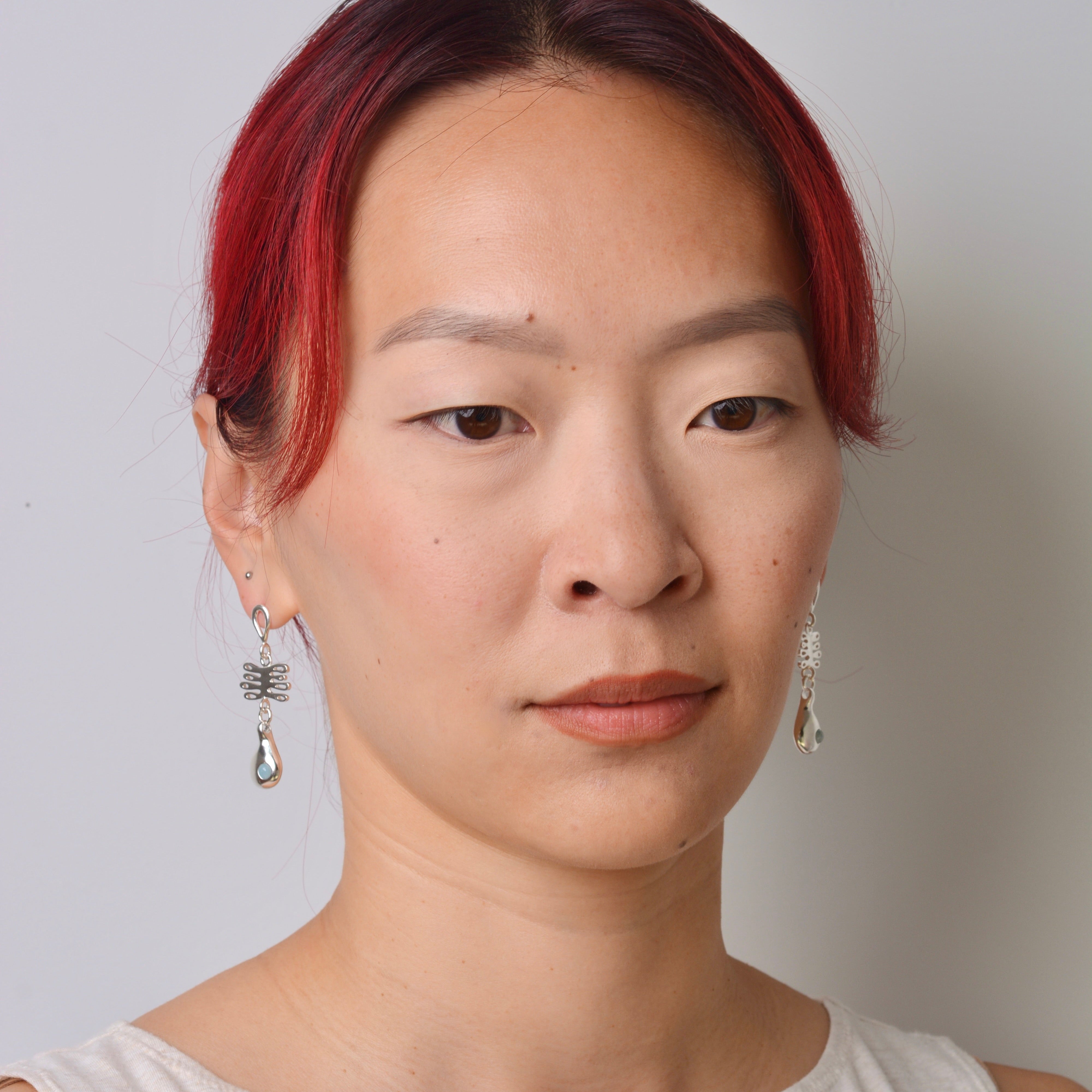 Person with red hair wearing silver dangle earrings against a light-colored top against a plain background