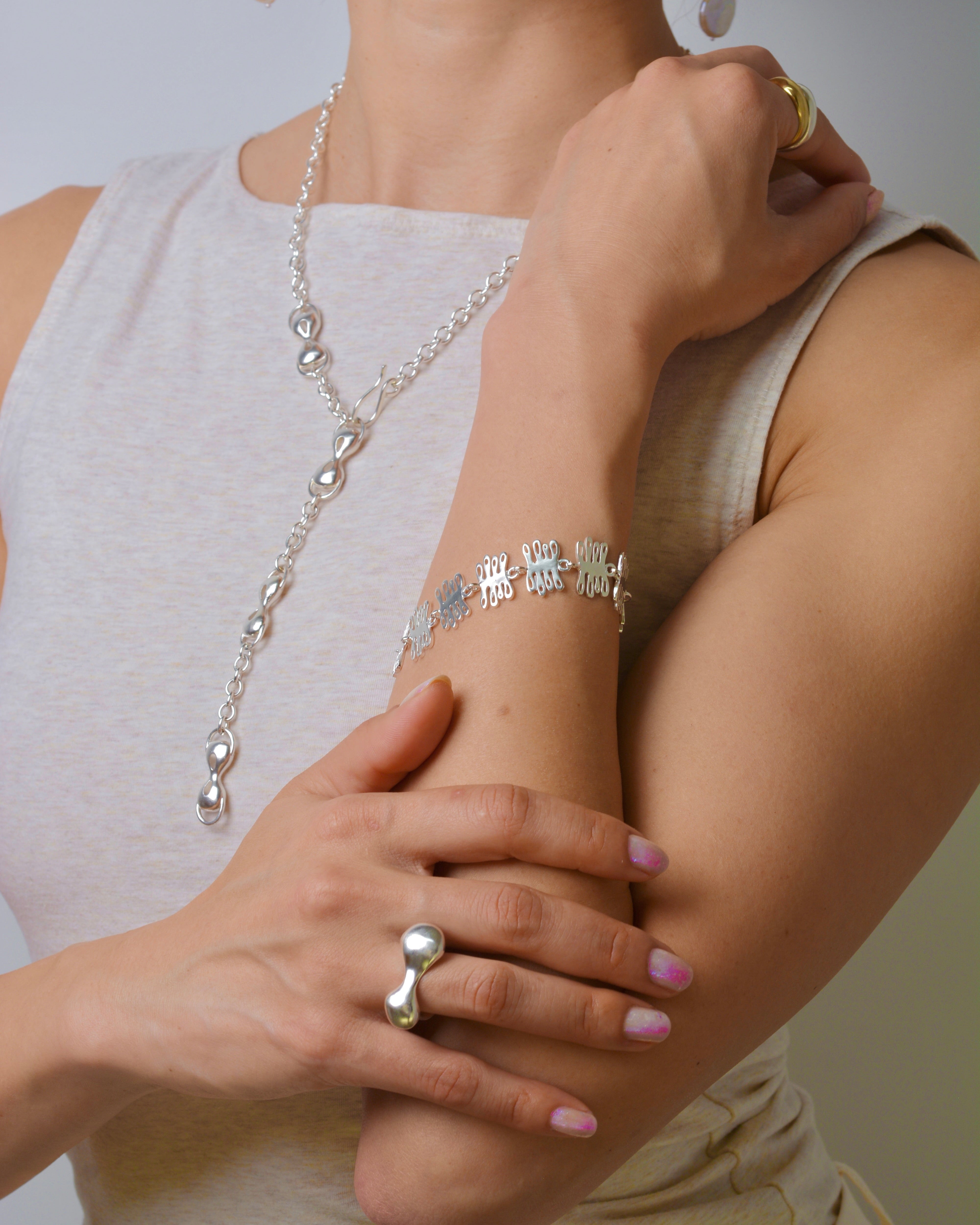 Person wearing silver jewelry including a long necklace, plant inspired bracelet, and organic rings on a neutral background