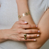 Close-up of a person wearing gold colored plant inspired jewelry on their wrist and organic shaped ring on their finger, with a neutral background.