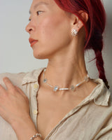 Woman wearing silver jewelry including a pearl and plant-shaped charm necklace and earrings against a neutral background