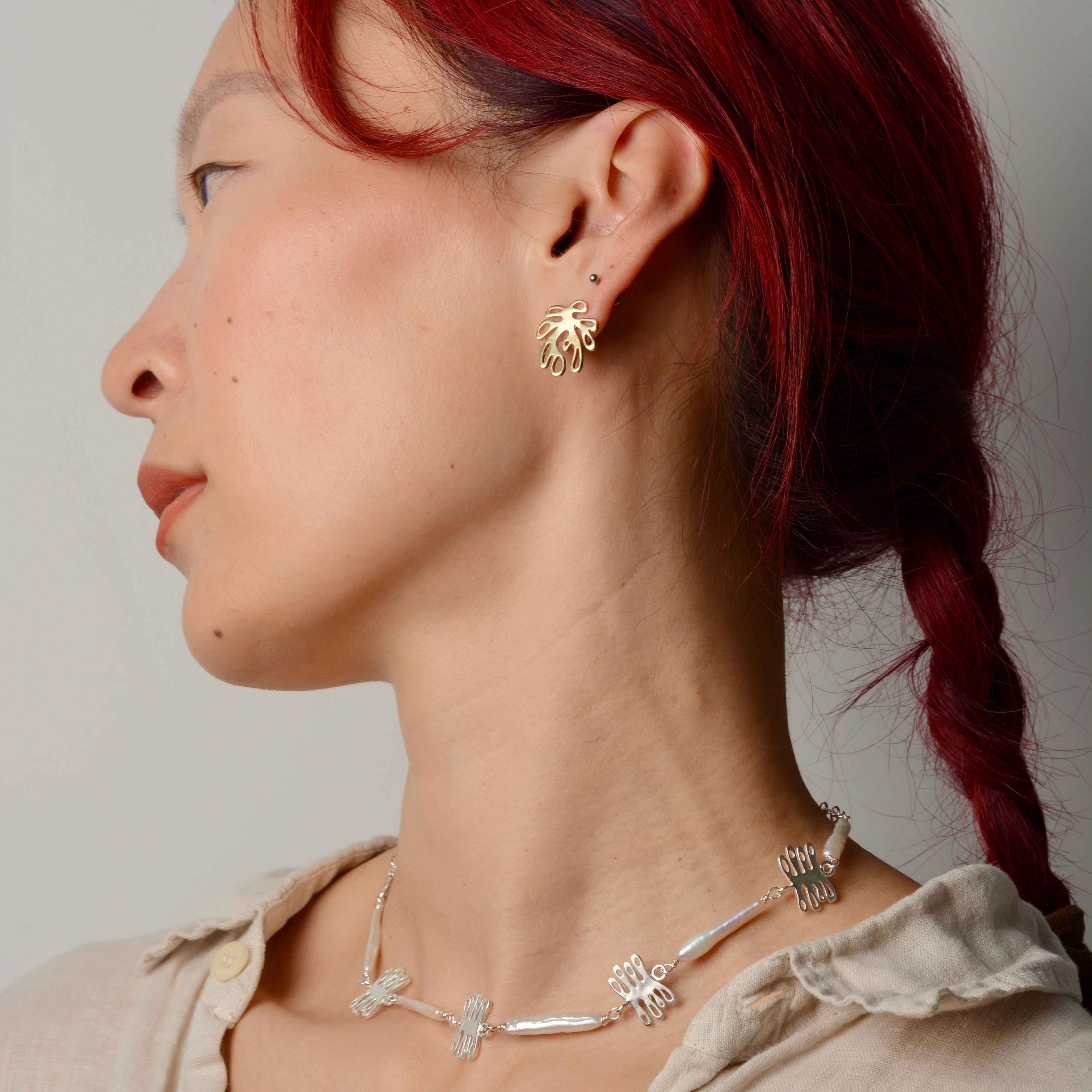 Woman with red hair wearing gold plant shaped earrings and a silver and pearl necklace against a plain background