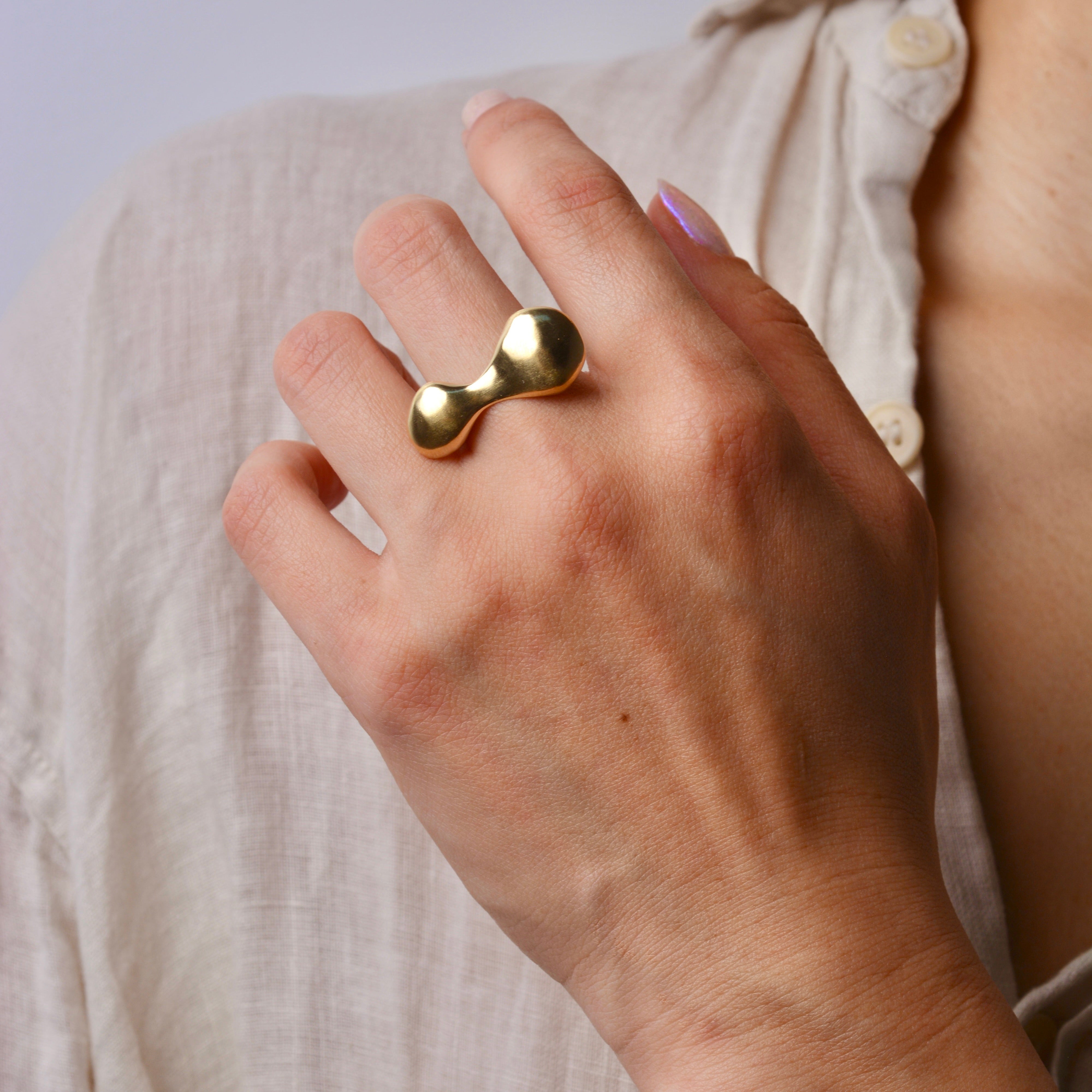Hand wearing a gold blob shaped ring with a plain background