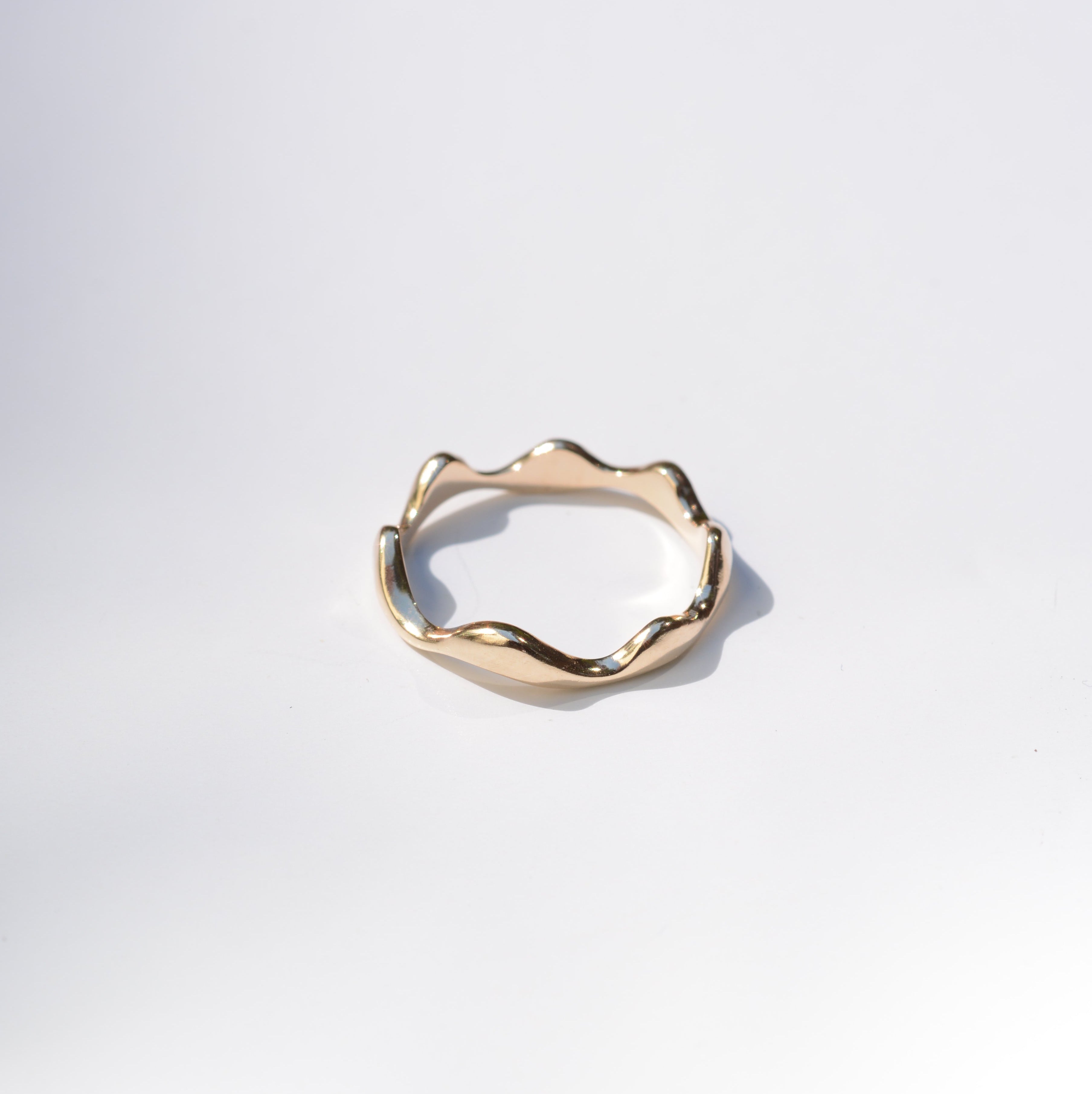 A single gold ring with an organic, wavy, undulating band design, photographed on a clean white background with a soft shadow.