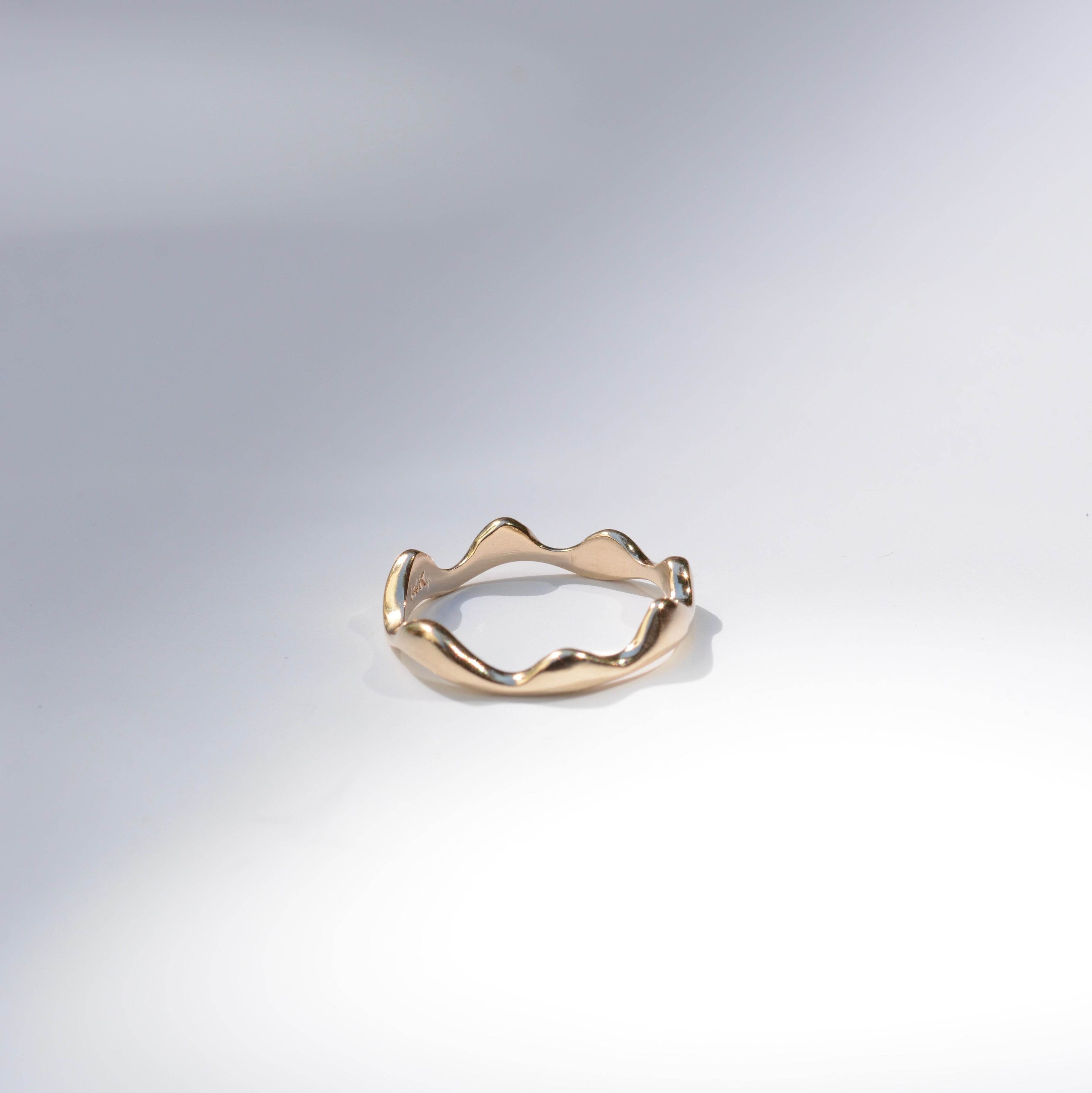 A single gold ring with an organic, wavy, undulating band design, photographed on a clean white studio background with soft shadows.