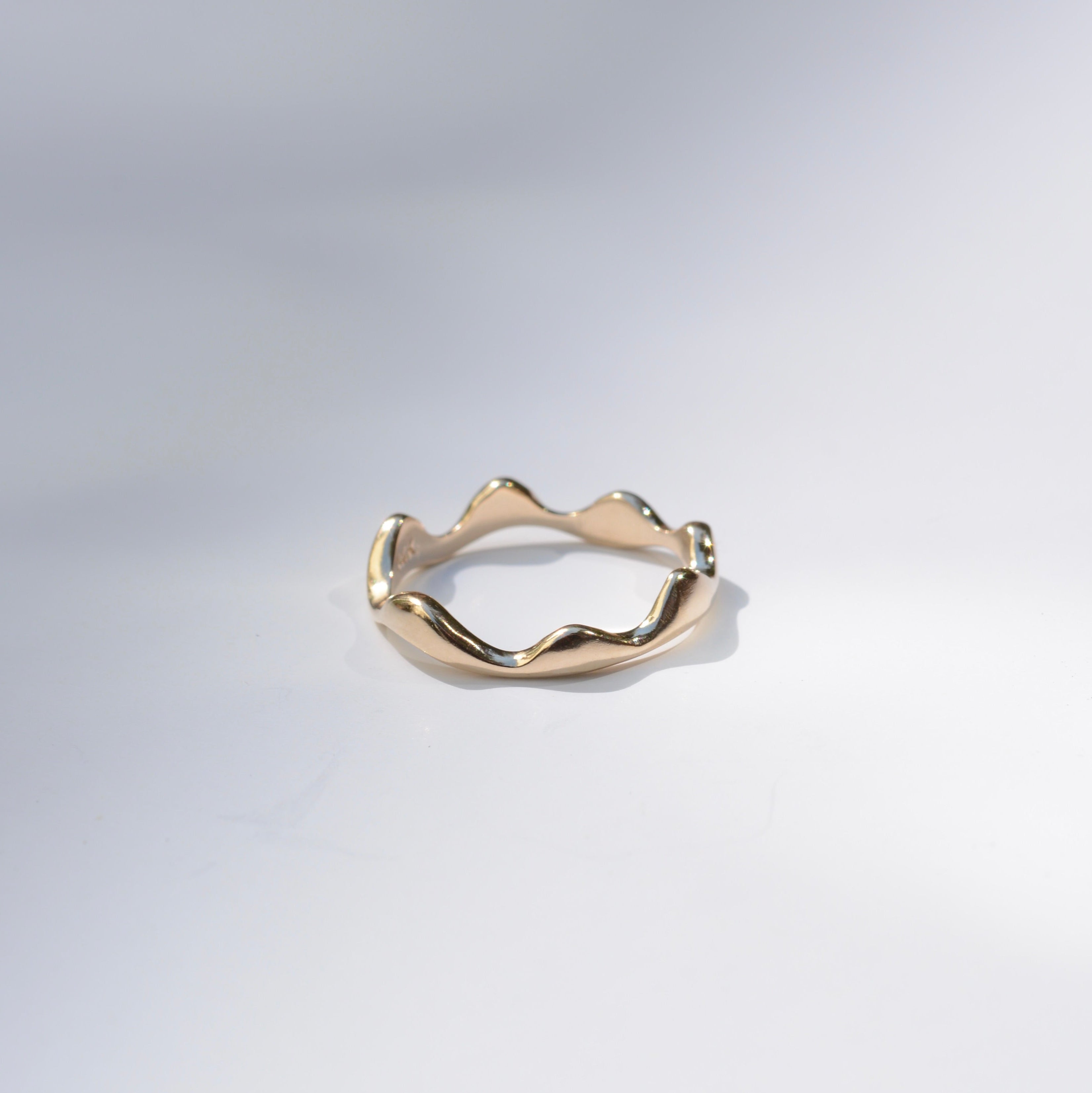 A gold ring with an organic, wavy, undulating band, photographed on a clean white studio background.