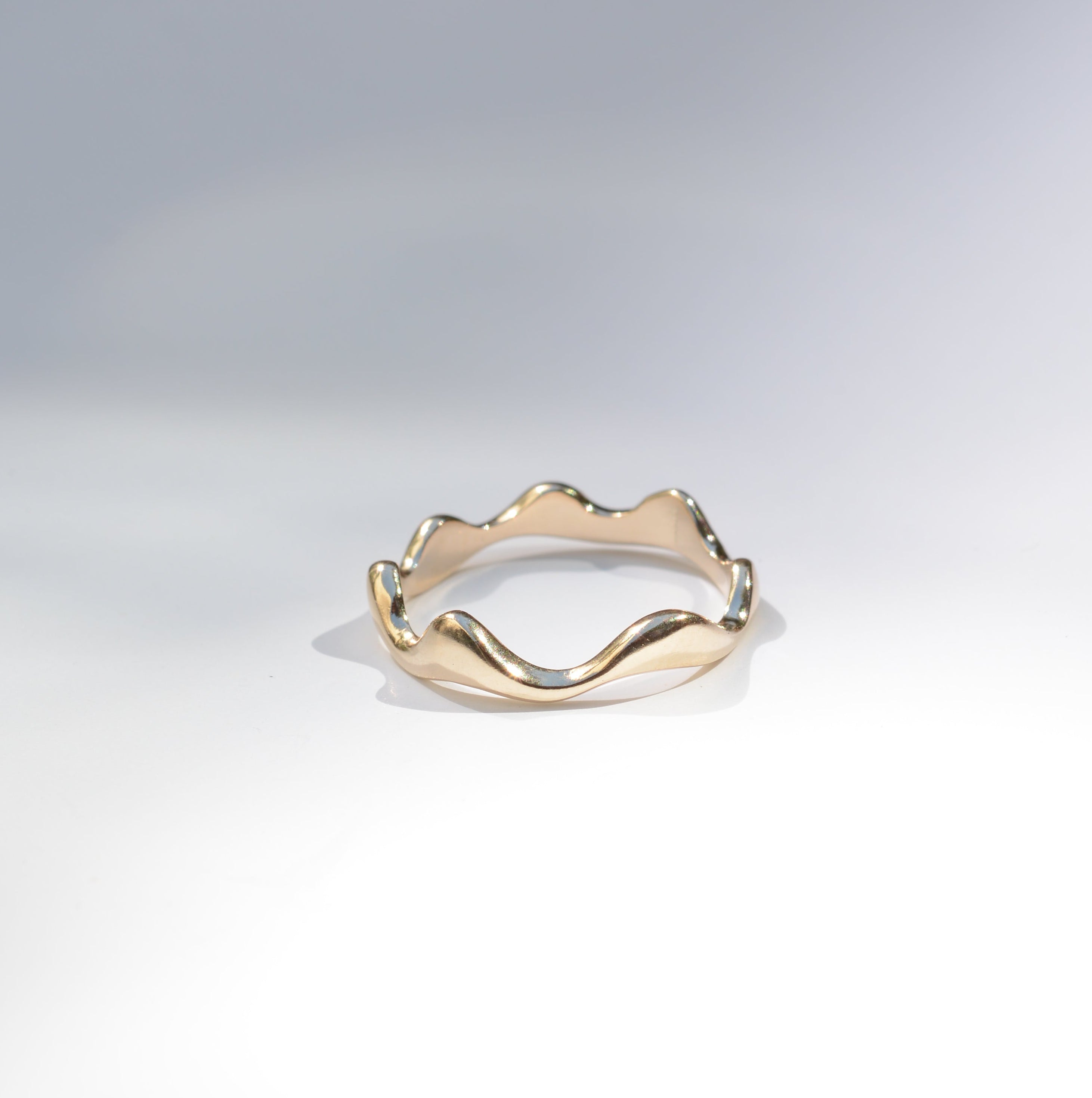 A gold wavy band ring with a scalloped, undulating edge, photographed on a clean white surface with soft shadows.