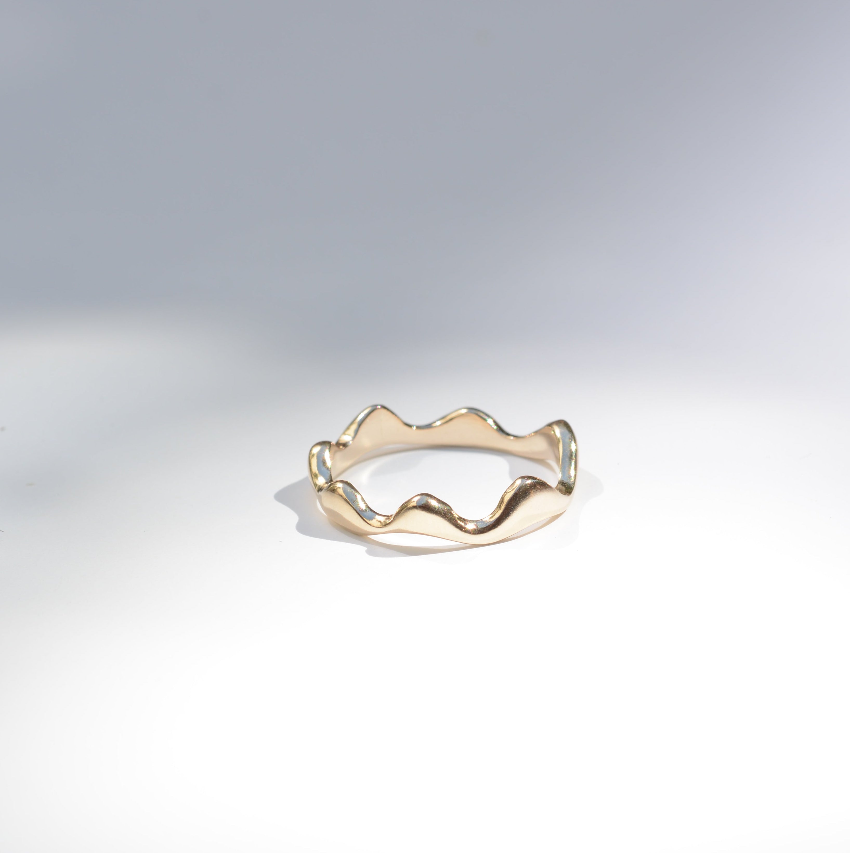 A thin gold ring with a wavy, undulating edge, photographed on a clean white surface with soft shadows.