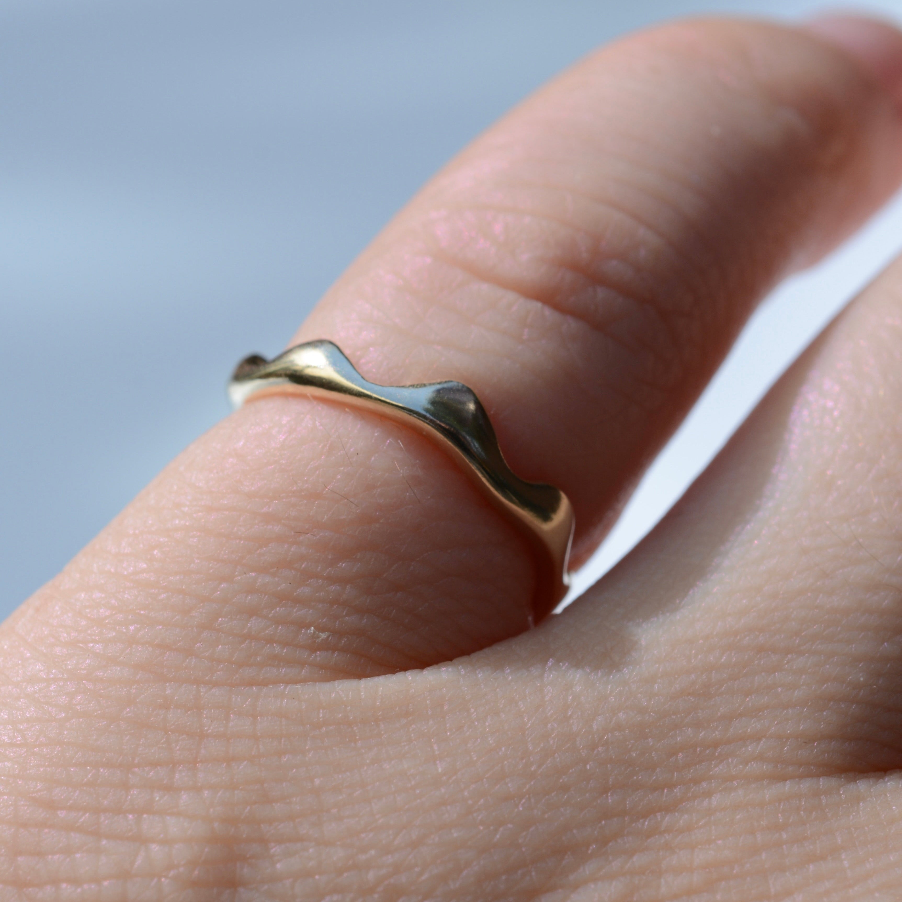 A close-up of a hand wearing a thin, wavy gold ring against a soft, light blue background.