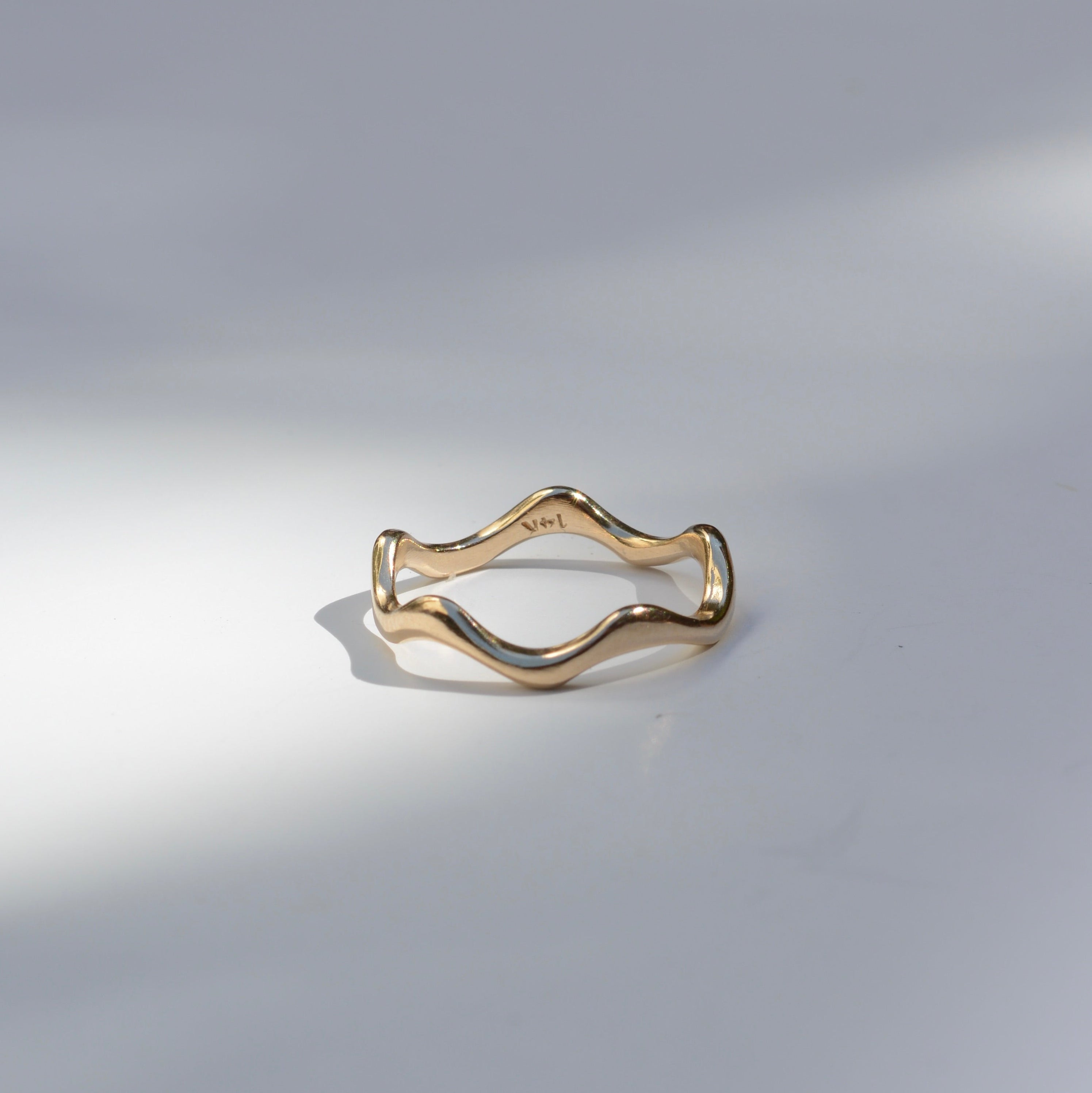 A thin wavy gold ring band photographed on a light gray surface with soft shadows.