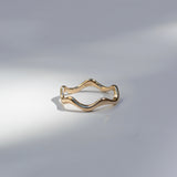 A thin wavy gold ring band photographed on a light gray surface with soft shadows.