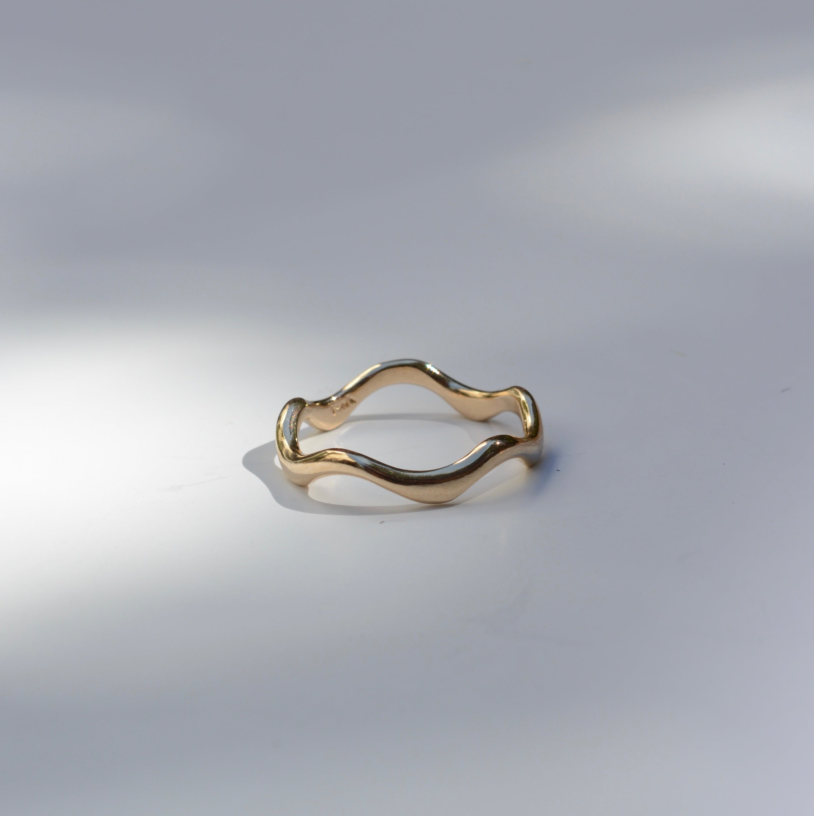 A single gold ring with a wavy, undulating band, photographed on a light gray surface with soft shadows.