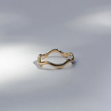 A single gold ring with a wavy, undulating band, photographed on a light gray surface with soft shadows.