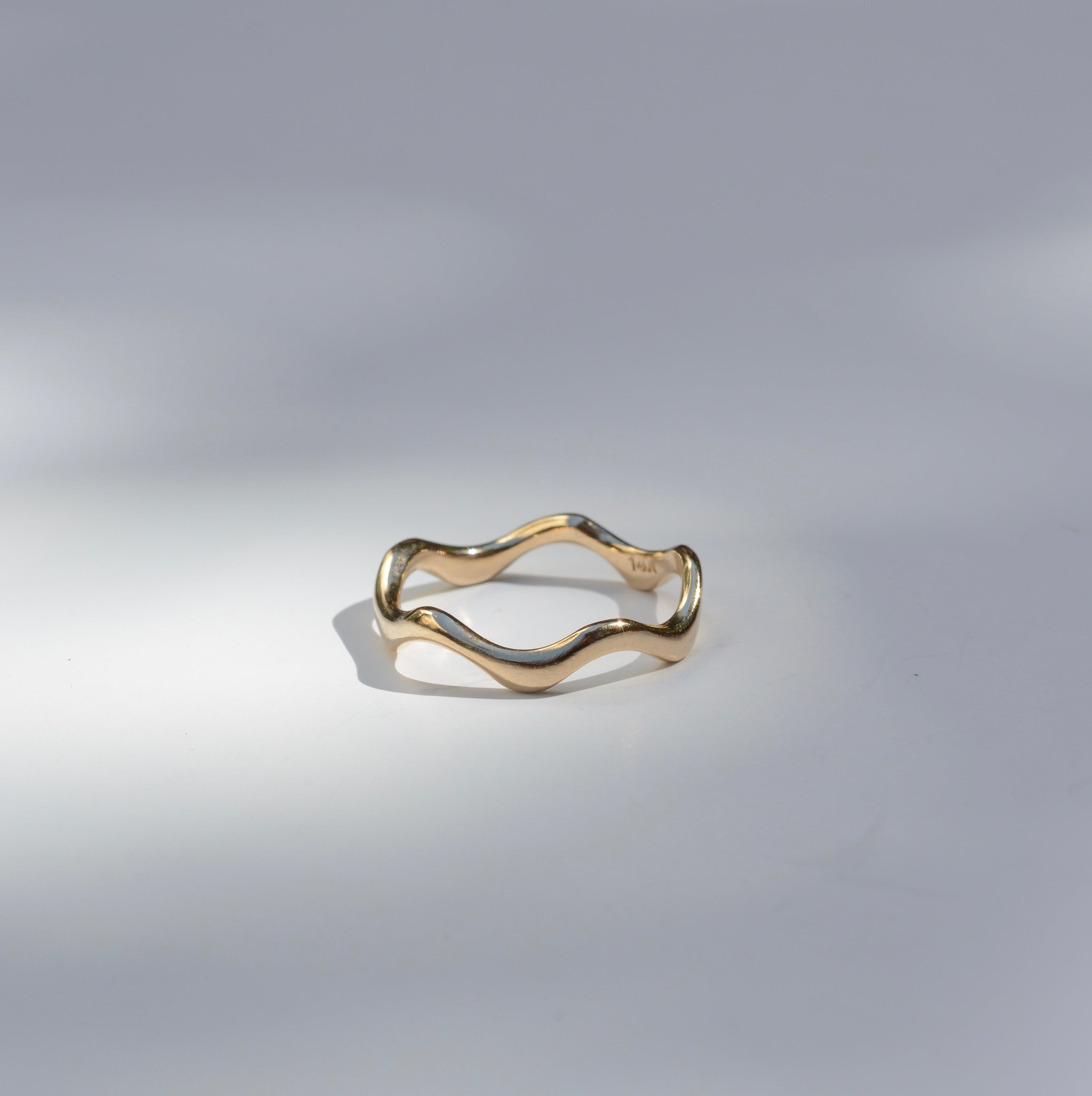 A thin wavy gold ring with a polished finish, photographed on a light gray surface with soft shadows.