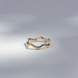 A thin wavy gold ring with a polished finish, photographed on a light gray surface with soft shadows.