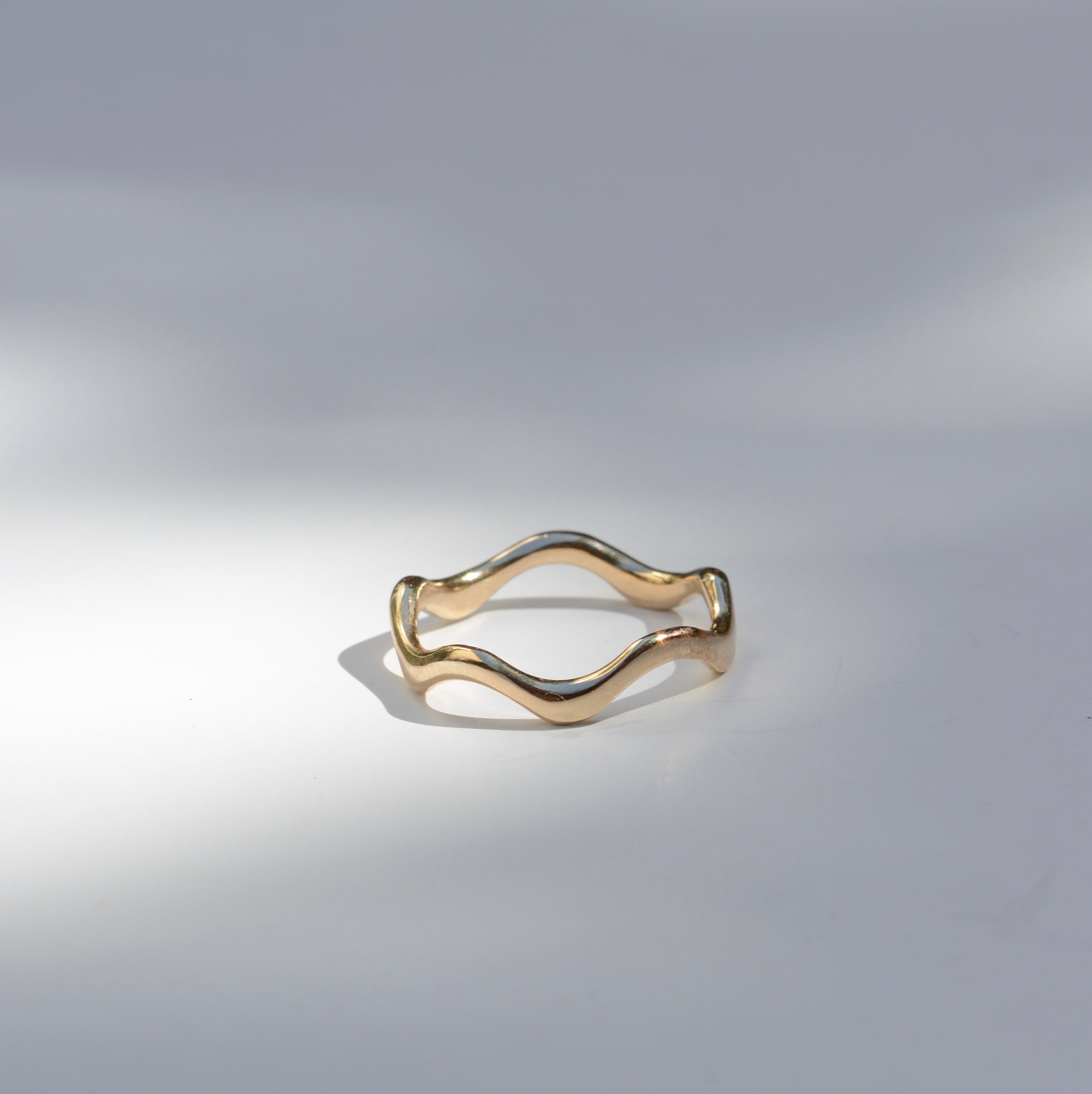 A thin wavy gold ring with a polished finish, displayed on a light gray surface with soft, artistic shadows.