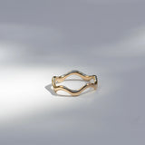 A thin wavy gold ring with a polished finish, displayed on a light gray surface with soft, artistic shadows.