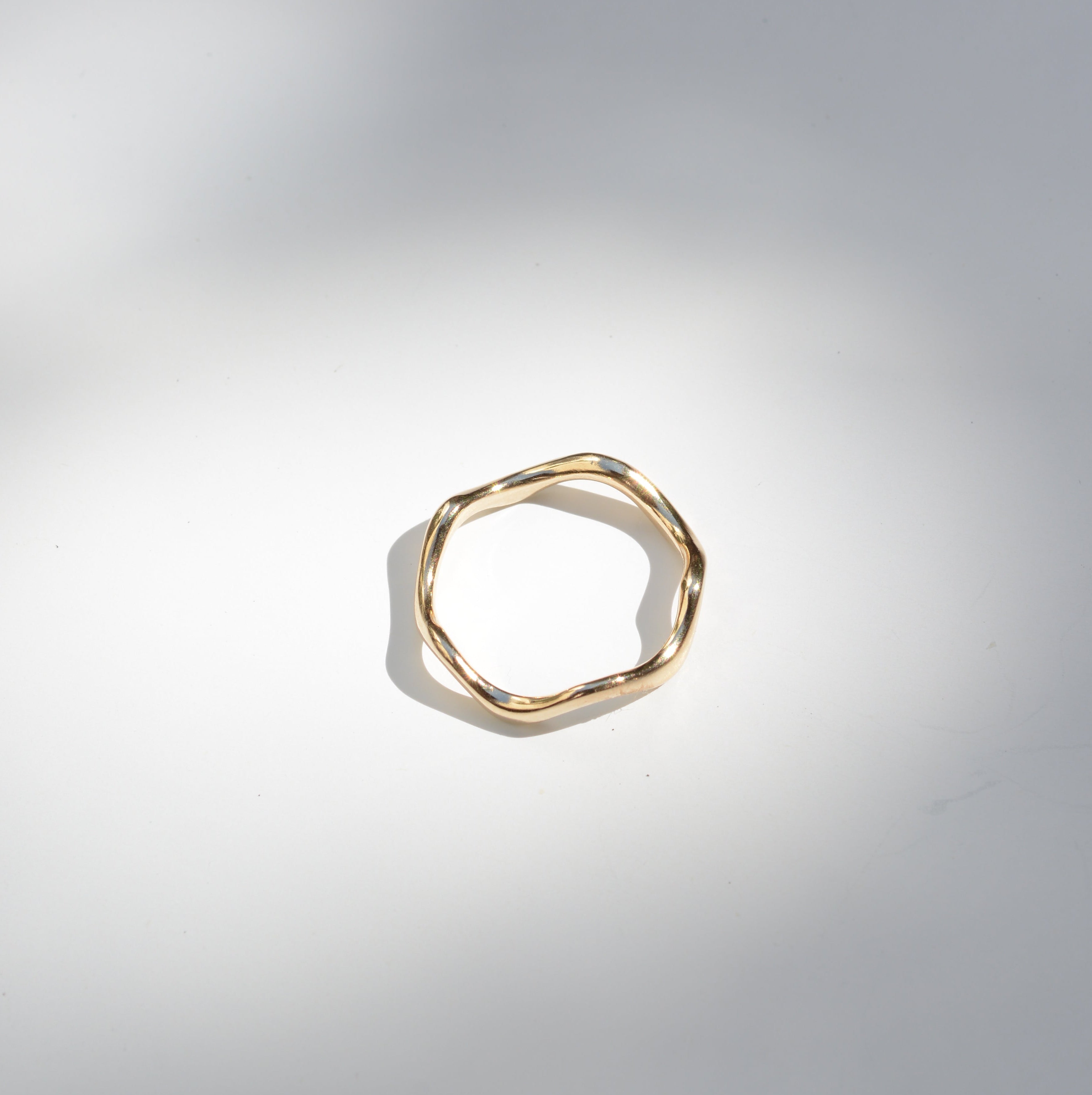 A single gold-toned ring with a textured, organic wavy shape, photographed on a light gray surface with soft shadows.
