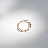A single gold-toned ring with a textured, organic wavy shape, photographed on a light gray surface with soft shadows.