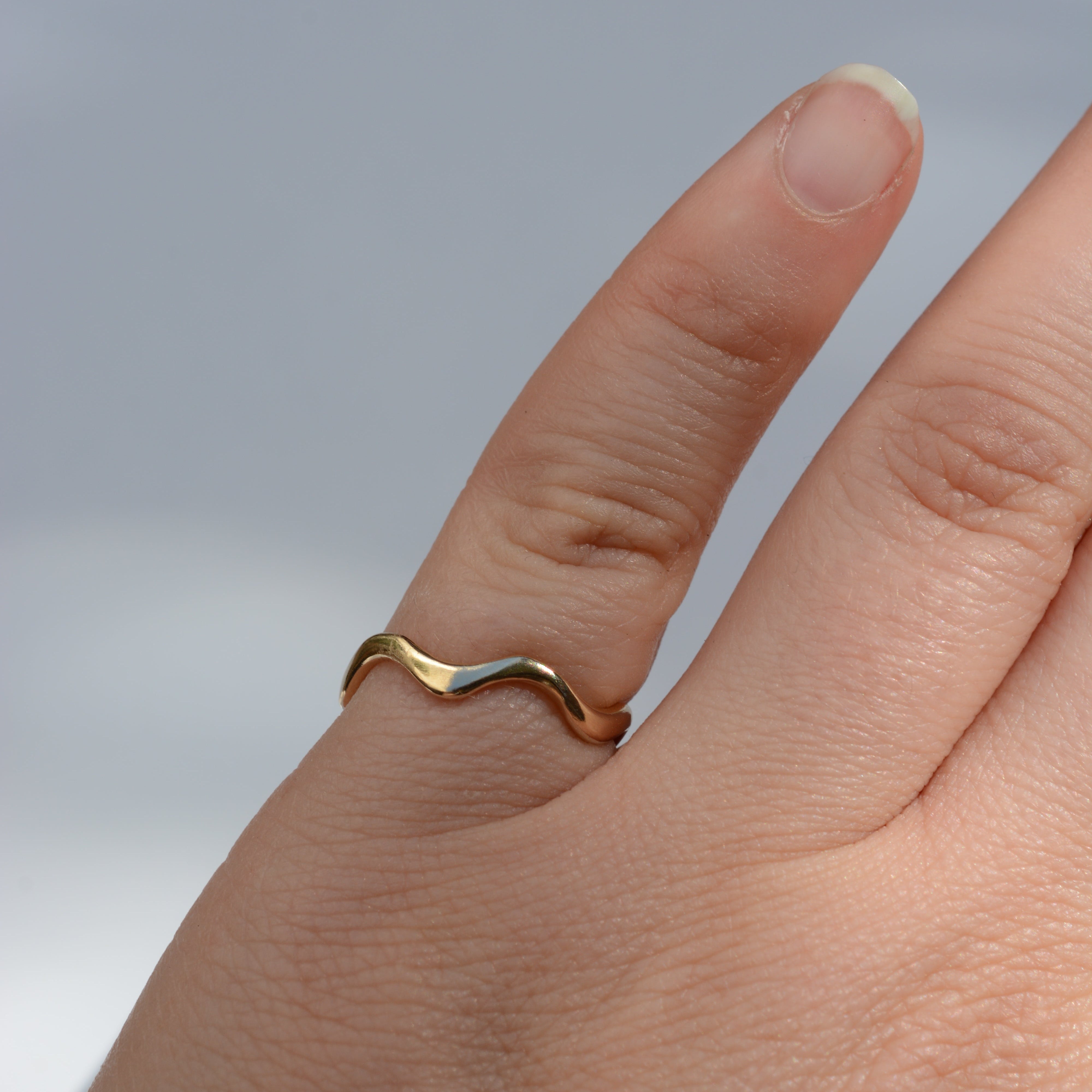 A close-up of a hand wearing a thin wavy band ring with a two-tone finish of yellow gold and silver, set against a soft, out-of-focus light gray background.