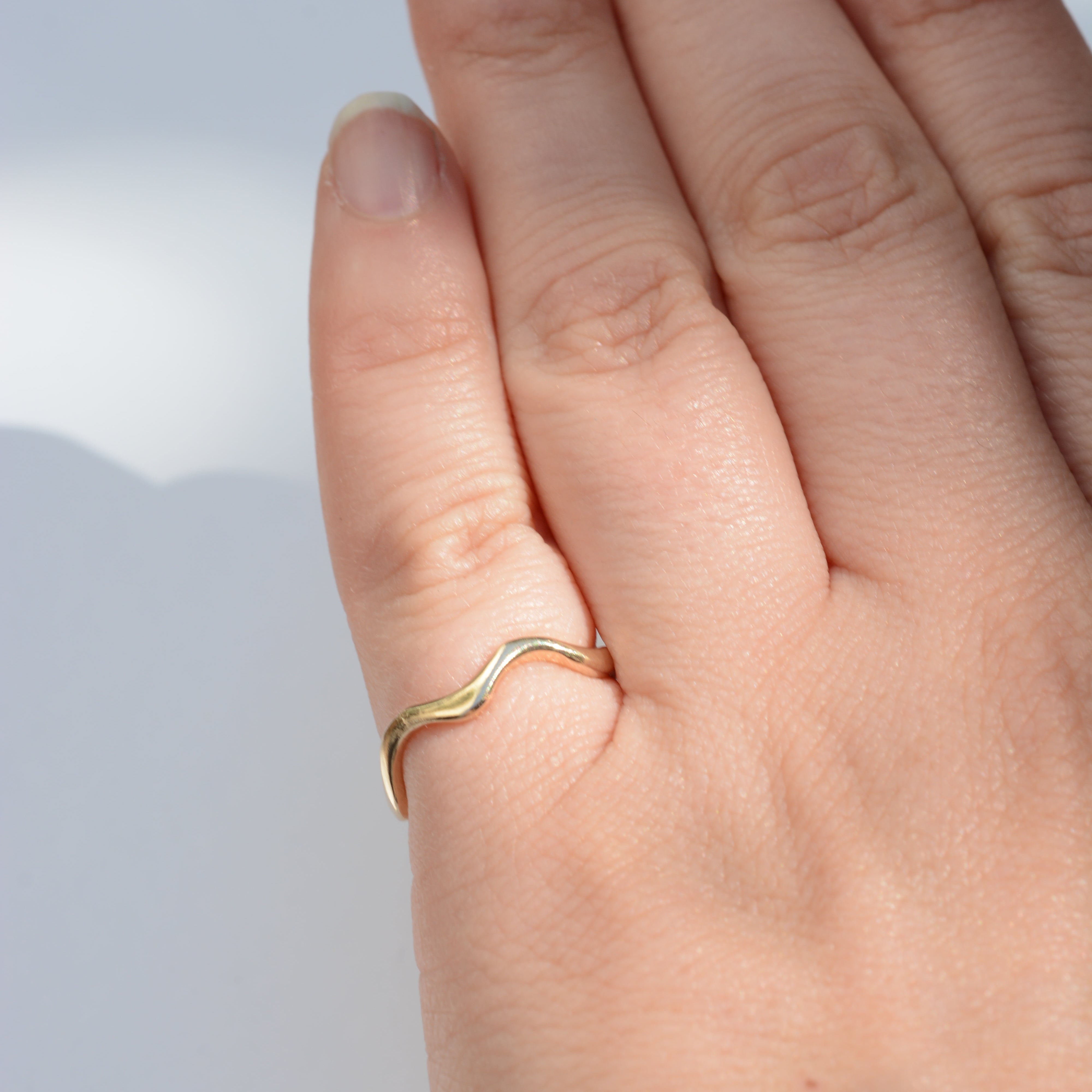 A close-up of a hand wearing a thin, wavy gold ring on one finger, set against a soft, light gray background.