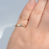 A close-up of a hand wearing a thin, polished gold ring with a small, abstract leaf-like protrusion on the band, set against a soft, light gray background.