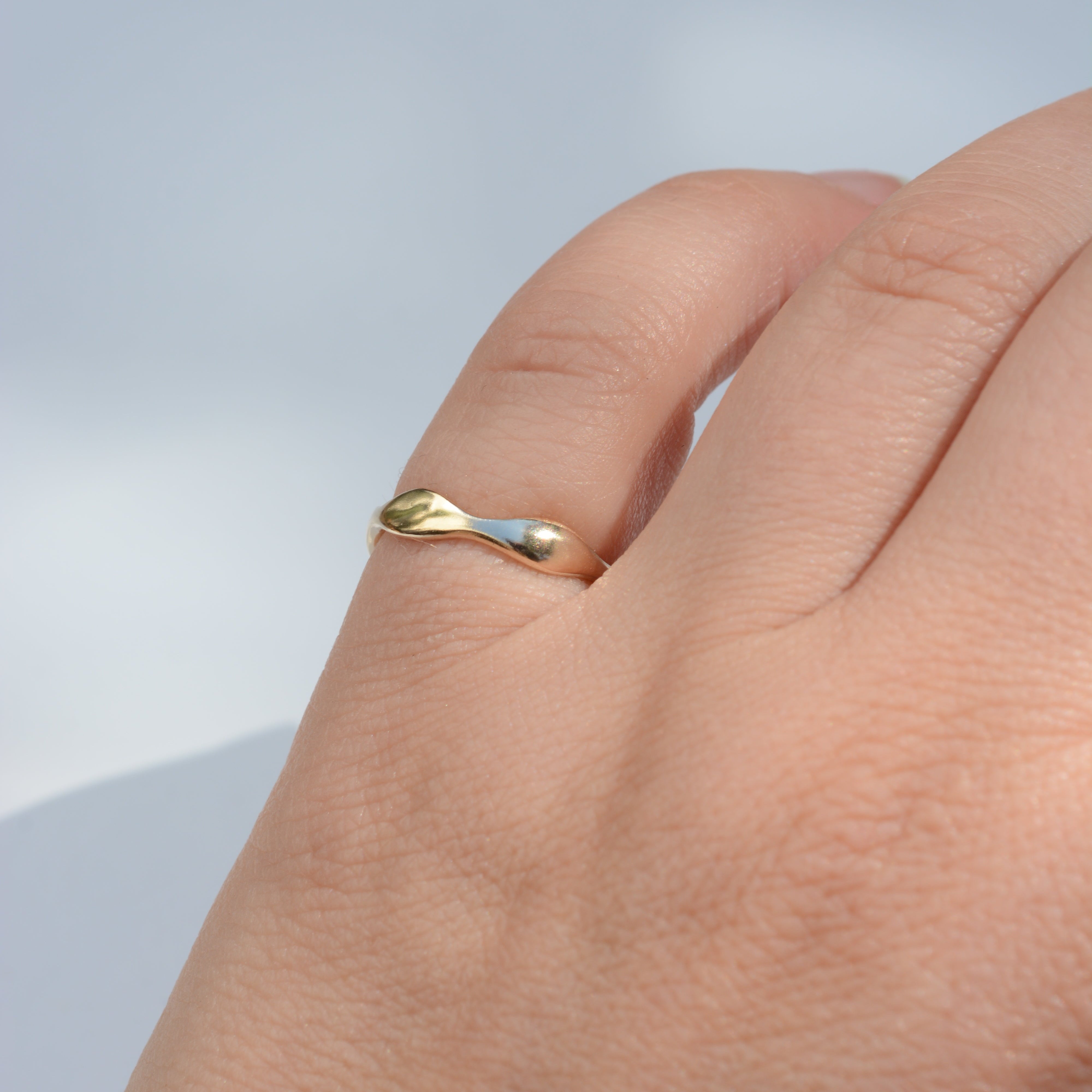 A close-up of a hand wearing a thin, polished gold ring with a unique wavy, organic shape against a soft, light blue-gray background.