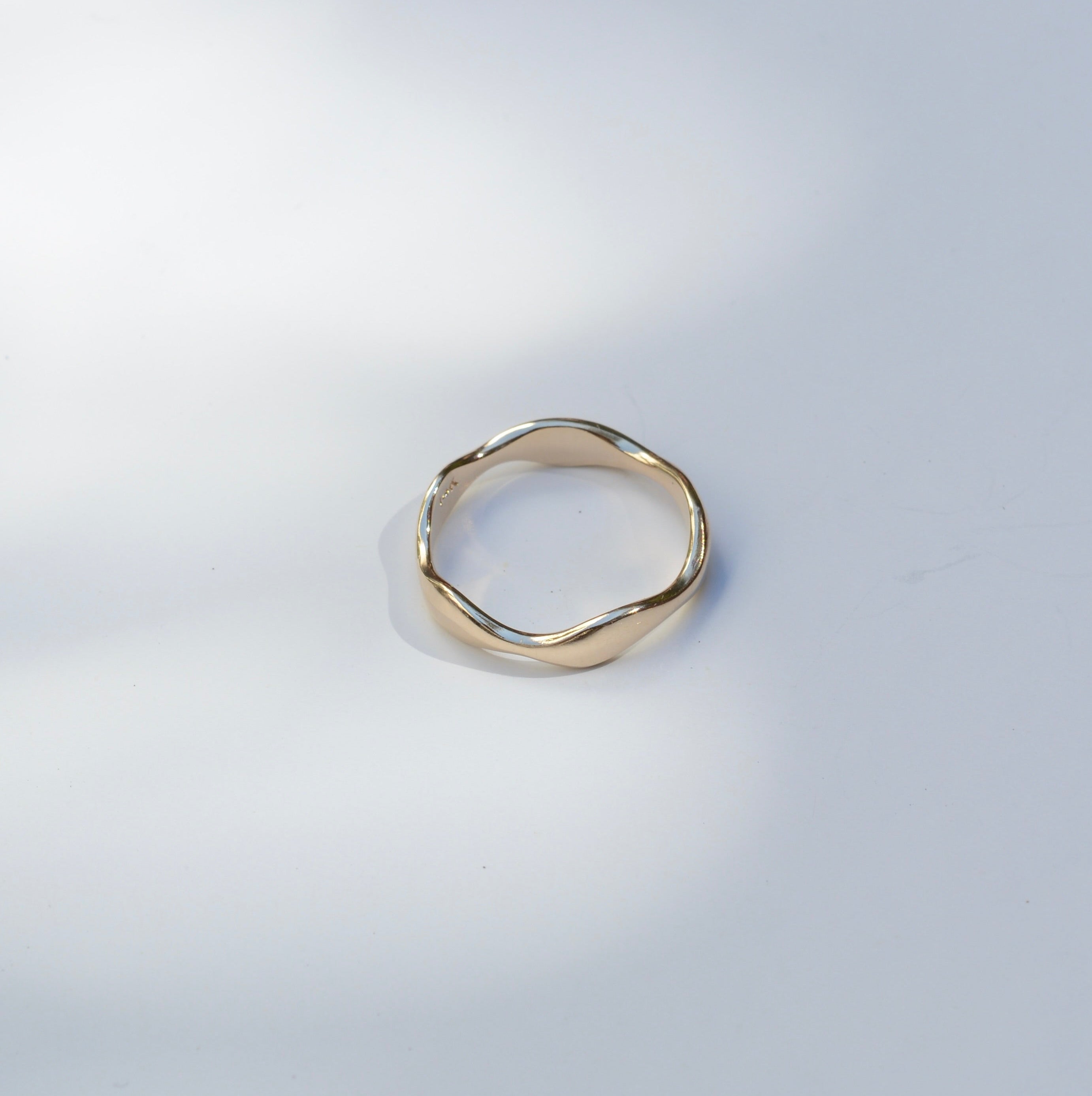 A single gold-toned ring with a wavy, organic textured band, photographed on a light gray surface with soft, dappled shadows.